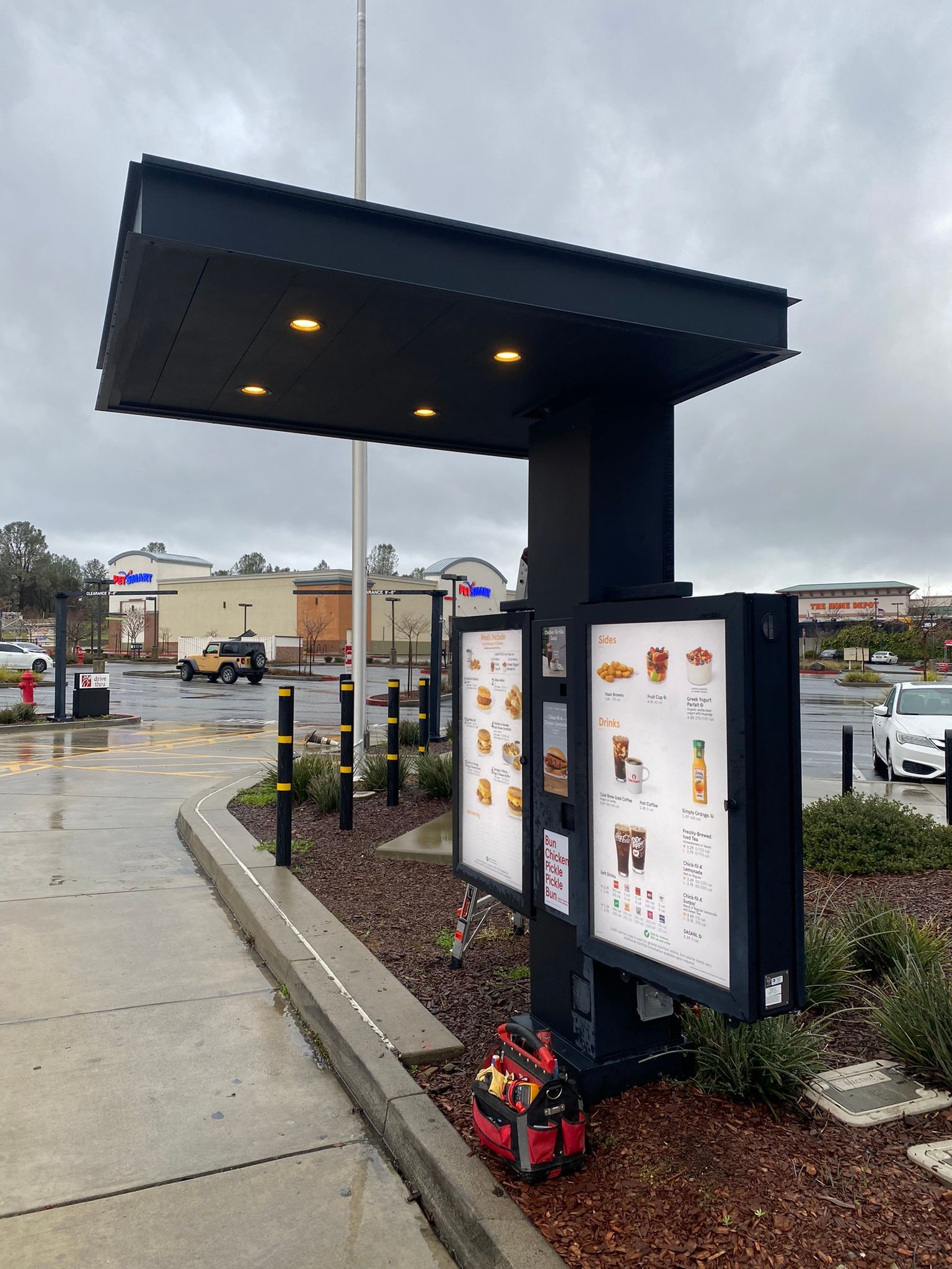 A black drive-thru menu board structure with overhead lighting stands in a paved lot on an overcast day.