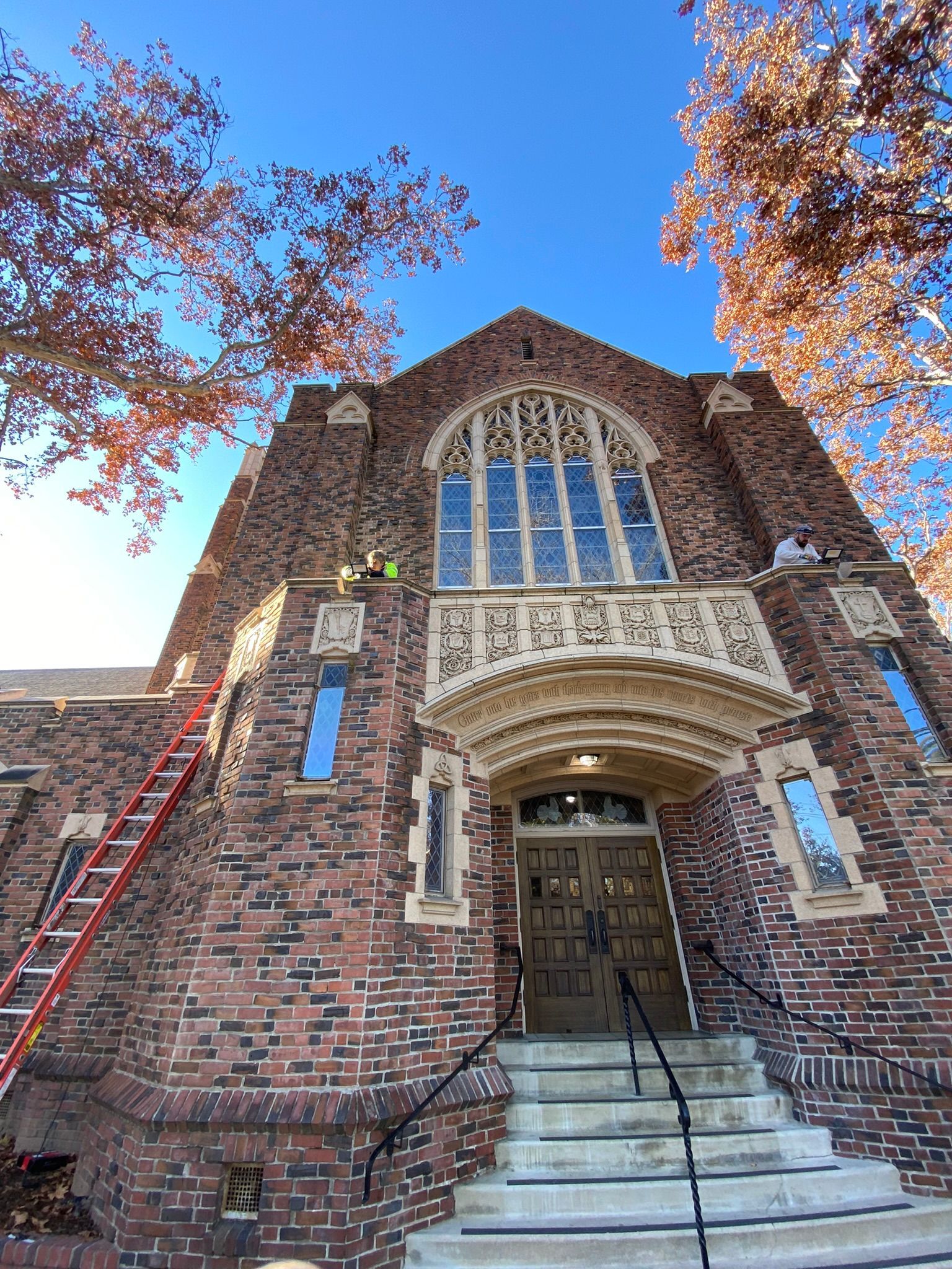 Low-angle view of a red brick church entrance with a tall arched window, stone detailing, and a ladder leaning against it.