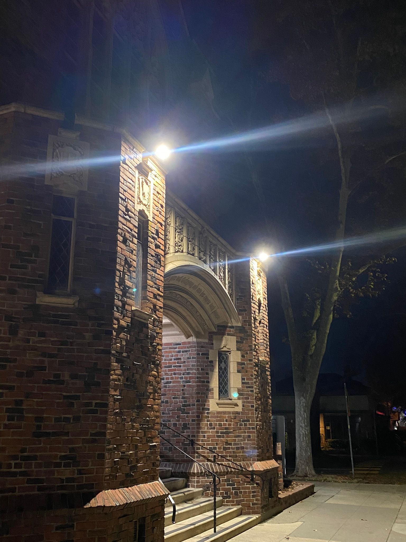 A brick archway at night, illuminated by bright spotlights, standing next to a large tree on a sidewalk.