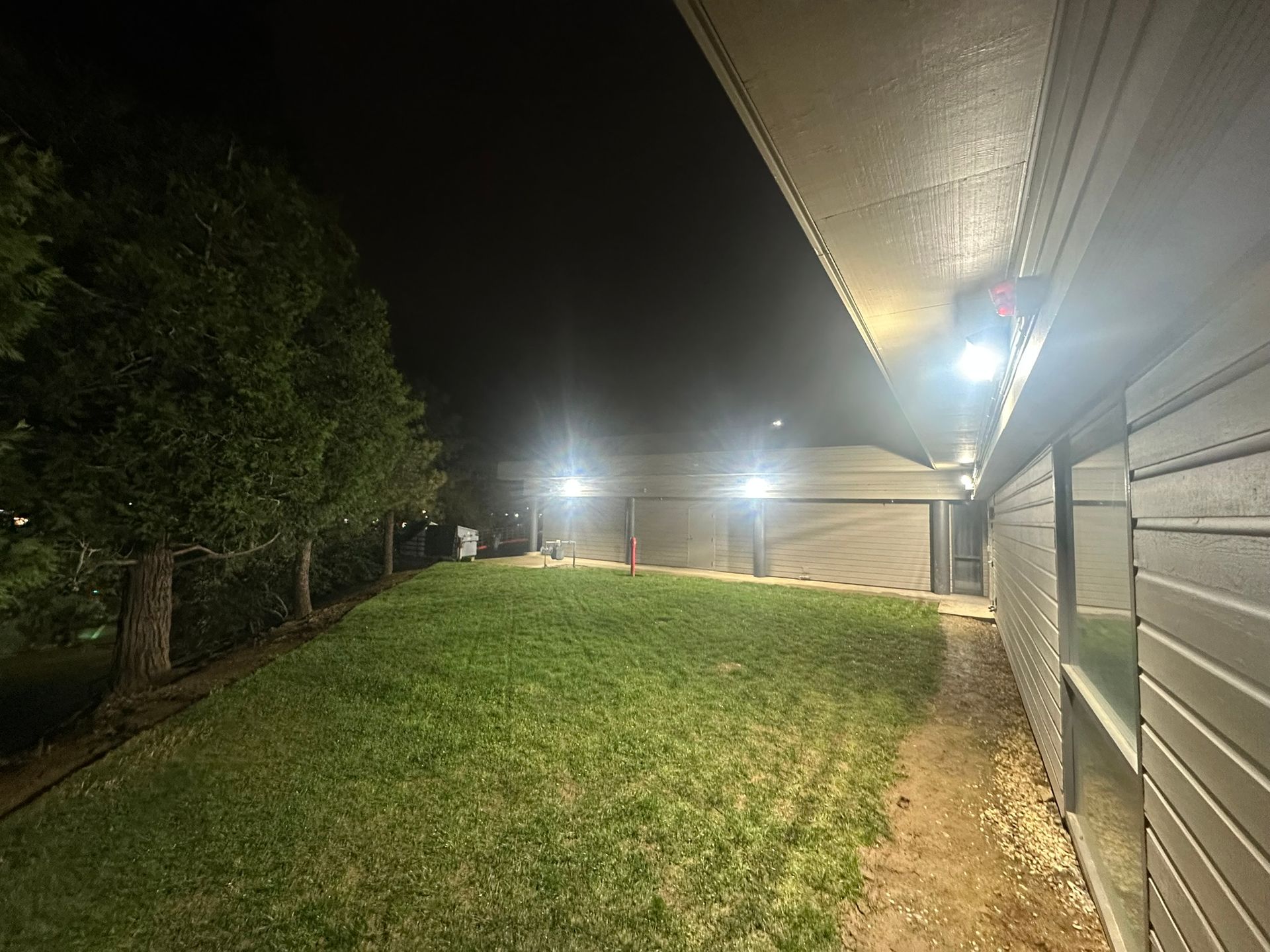 Night view of a residential side yard illuminated by bright exterior lights mounted under the roof eaves.