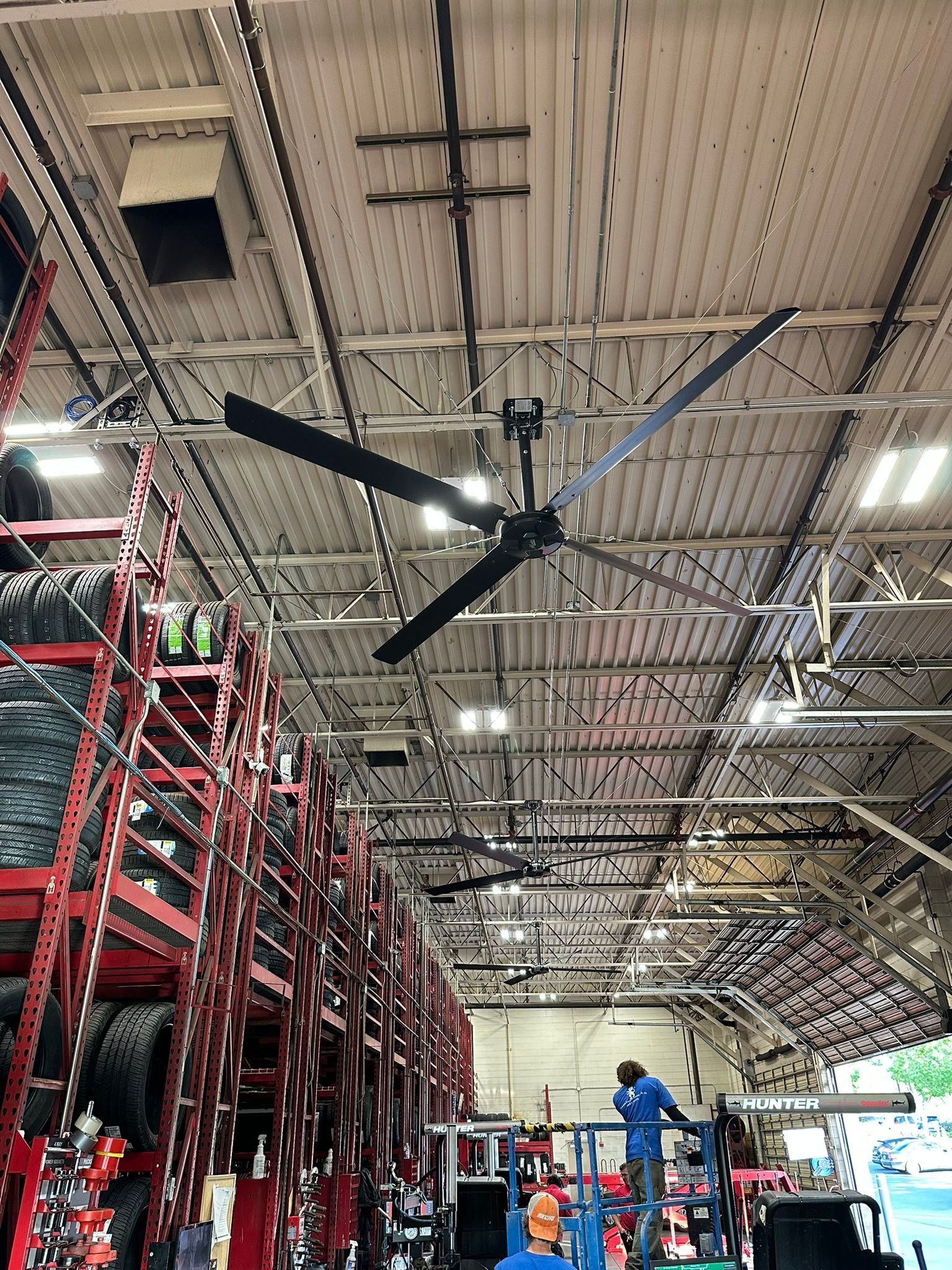 A large black industrial ceiling fan hangs in a warehouse with rows of red storage racks filled with tires.