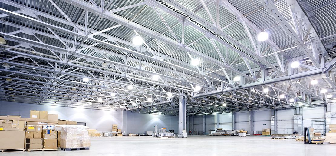 A high-ceilinged, industrial warehouse with bright lights and metal truss roofing, featuring pallets of stacked boxes.