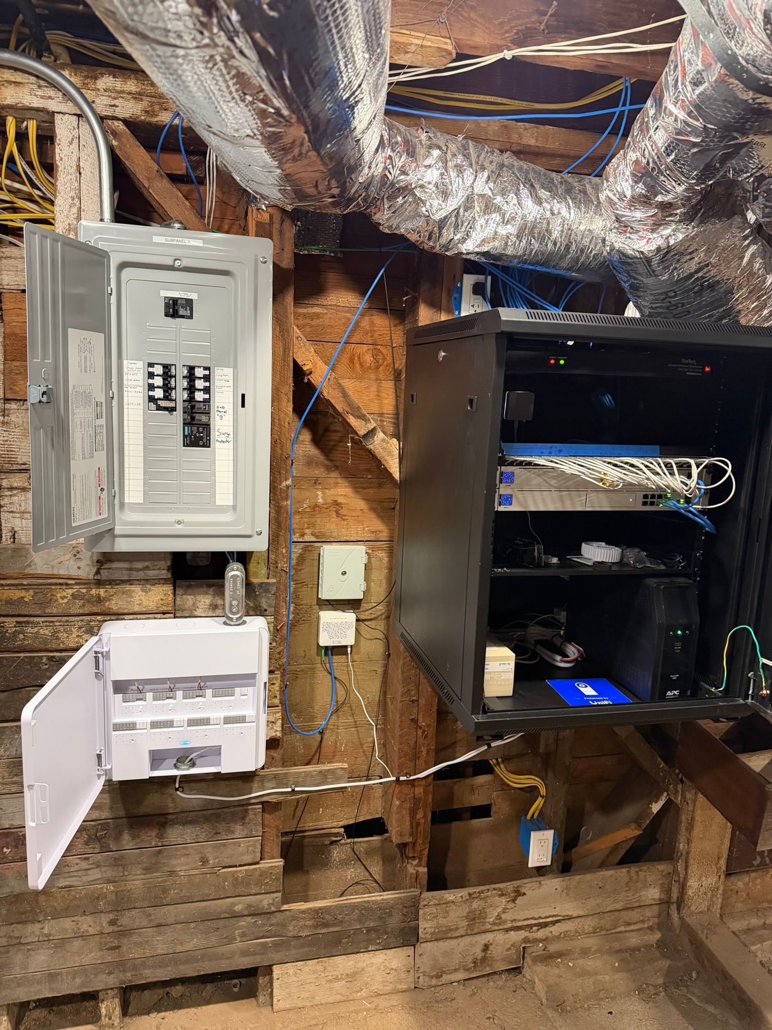 Electrical breaker panels and a black data rack with networking equipment mounted on an unfinished wooden wall.