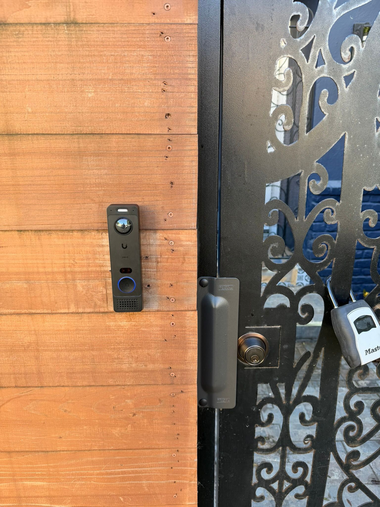 A black video doorbell mounted on a wooden post next to a decorative black metal gate with a keypad lock.