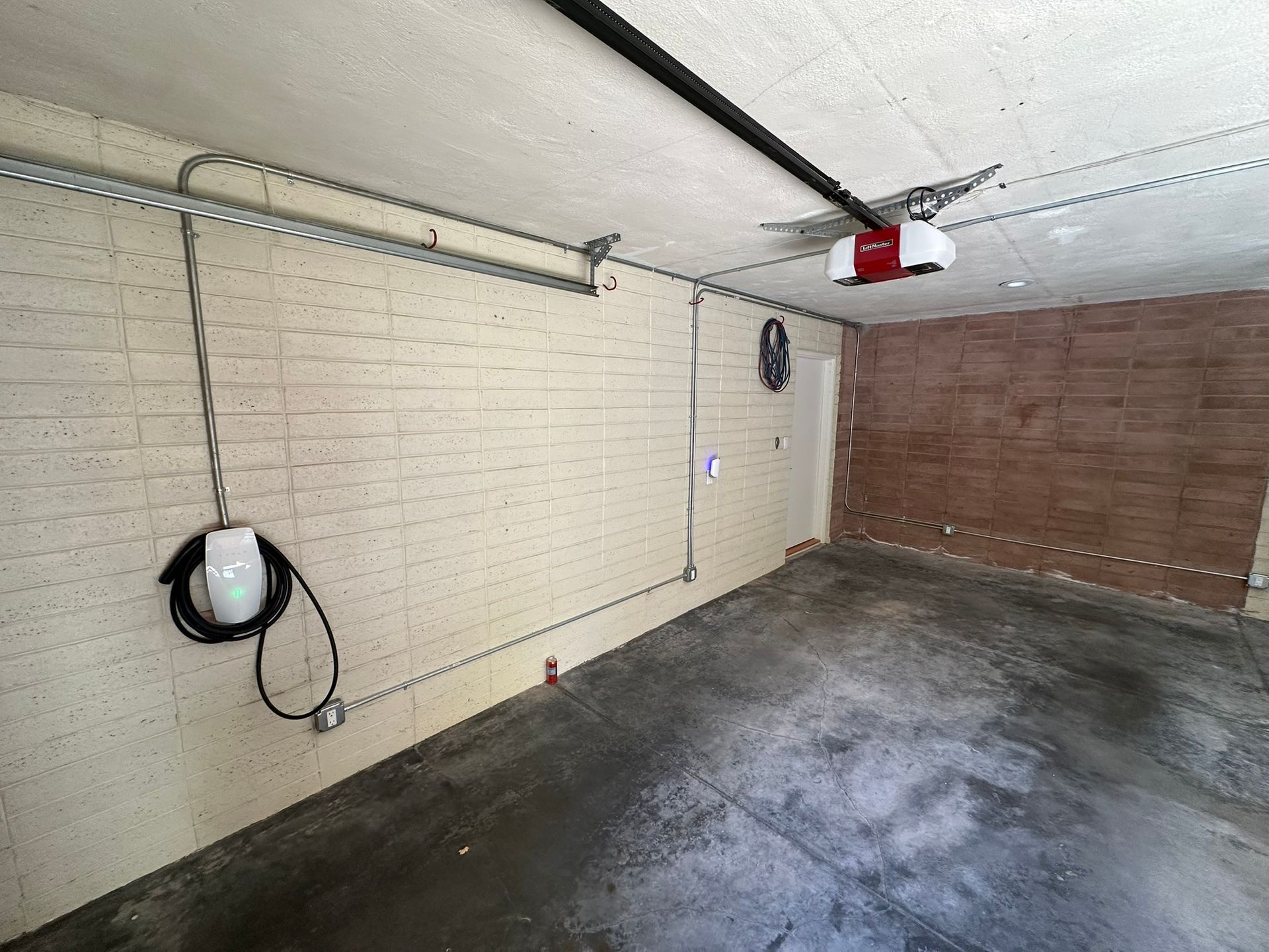 Empty garage with beige painted brick walls, a dark concrete floor, and an EV charging cable coiled on the wall.