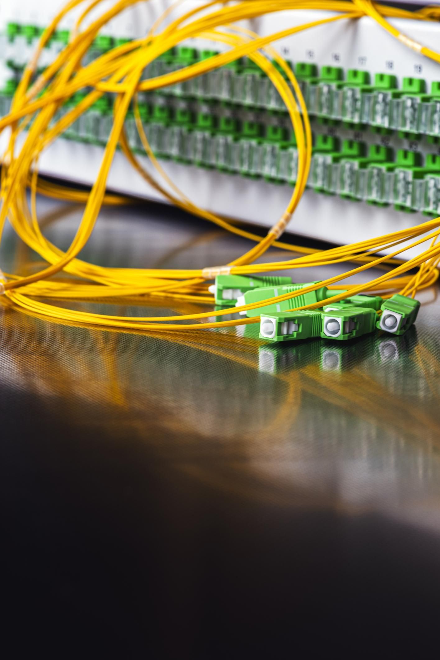 Green fiber optic connectors and yellow cables resting on a reflective surface in front of a patch panel.