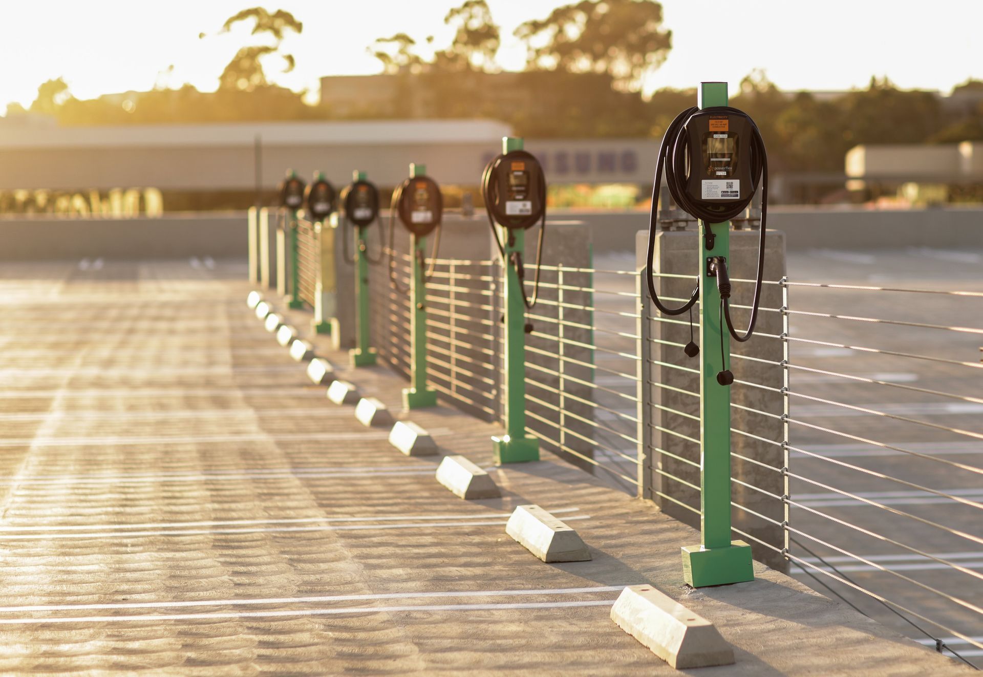 A row of green EV charging stations stands on a concrete rooftop parking deck at sunset, with a blurred building background.