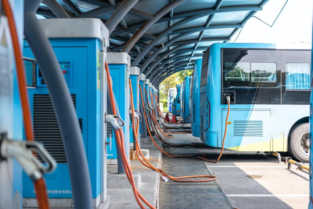 A row of blue electric buses parked under a metal canopy, connected to charging stations by orange cables.