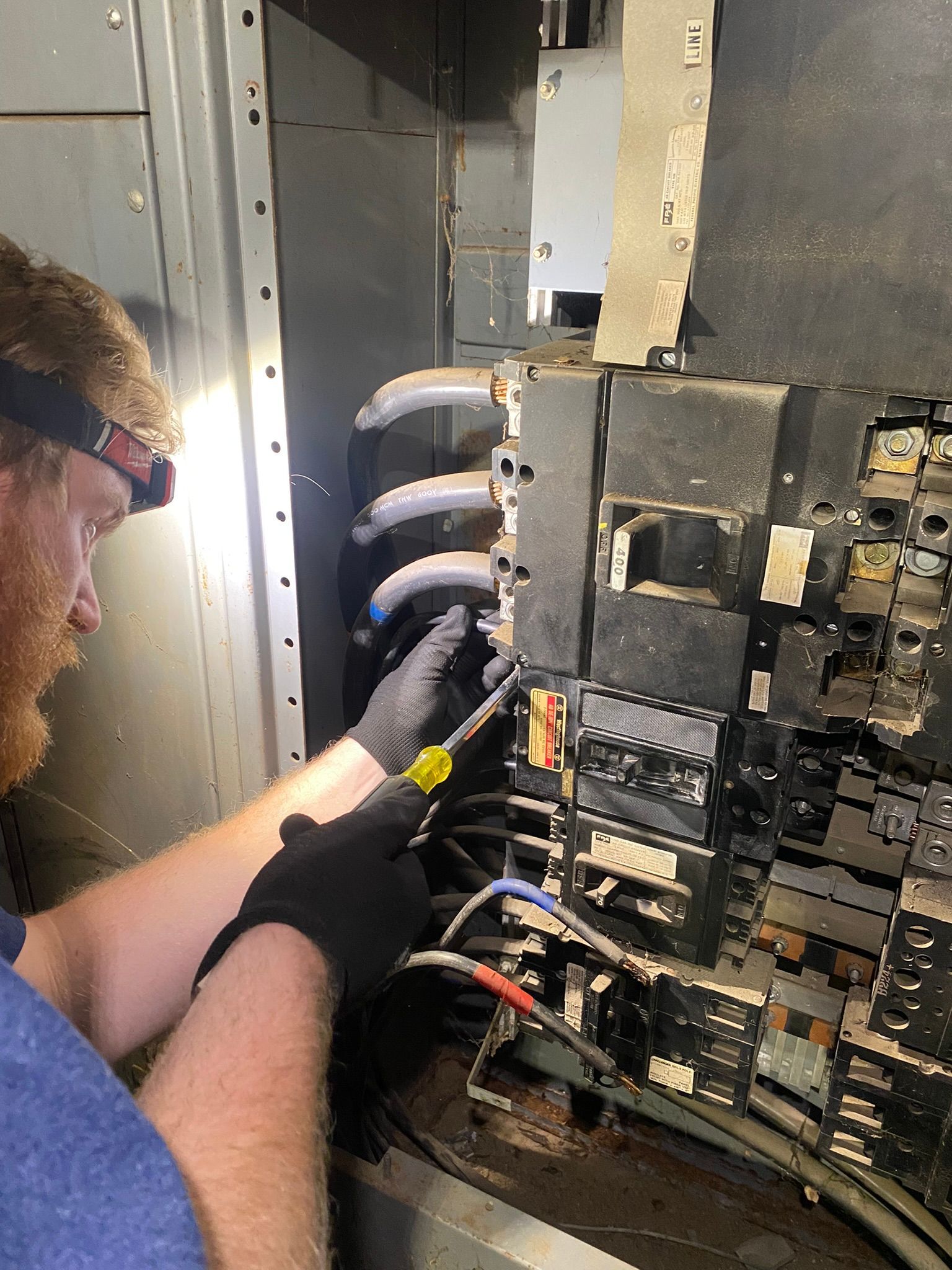 An electrician in protective gloves uses a screwdriver to work on a commercial electrical panel.