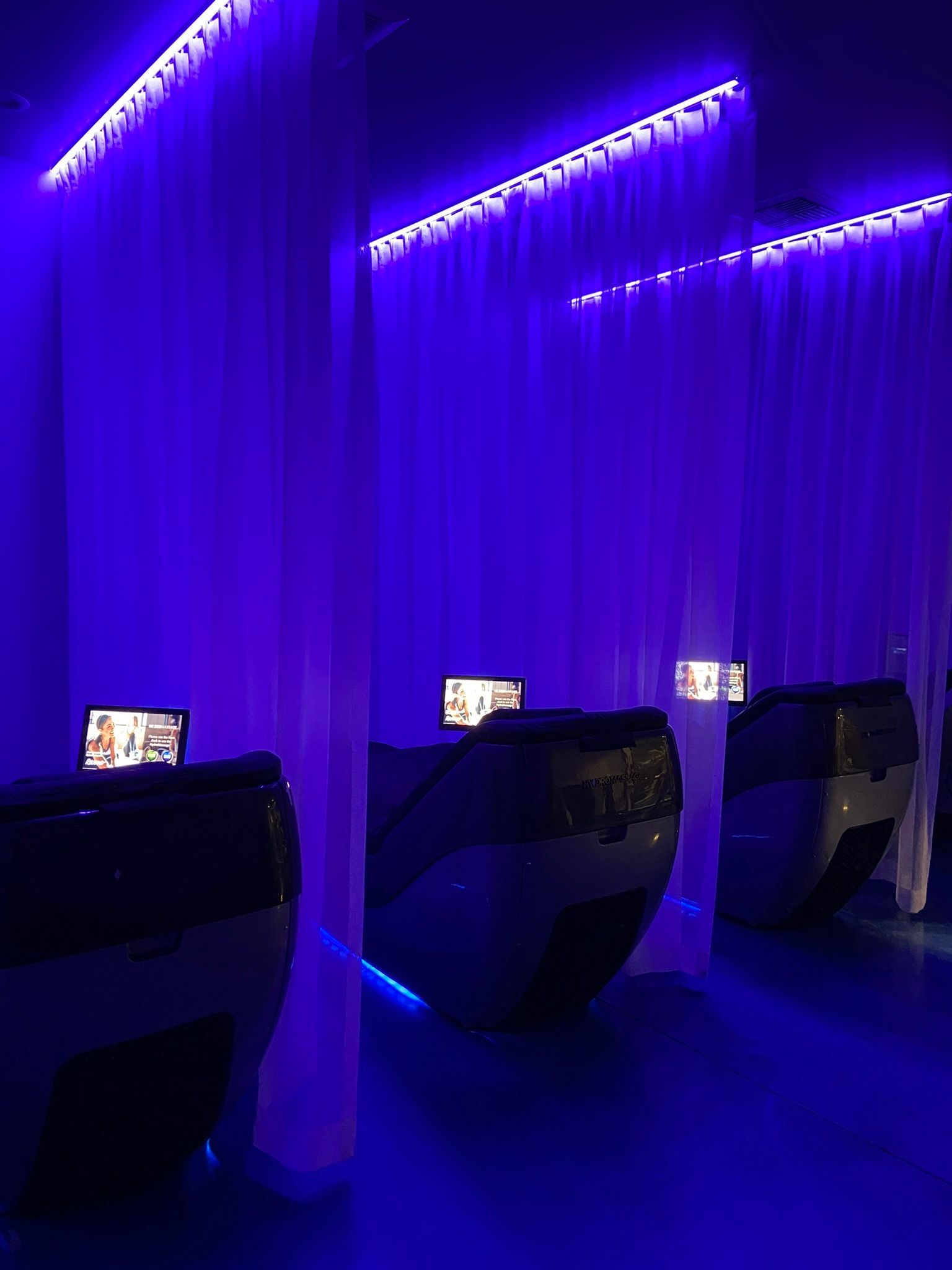 Three modern, individual privacy pods with glowing screens are separated by white curtains in a dimly lit, blue-lit room.