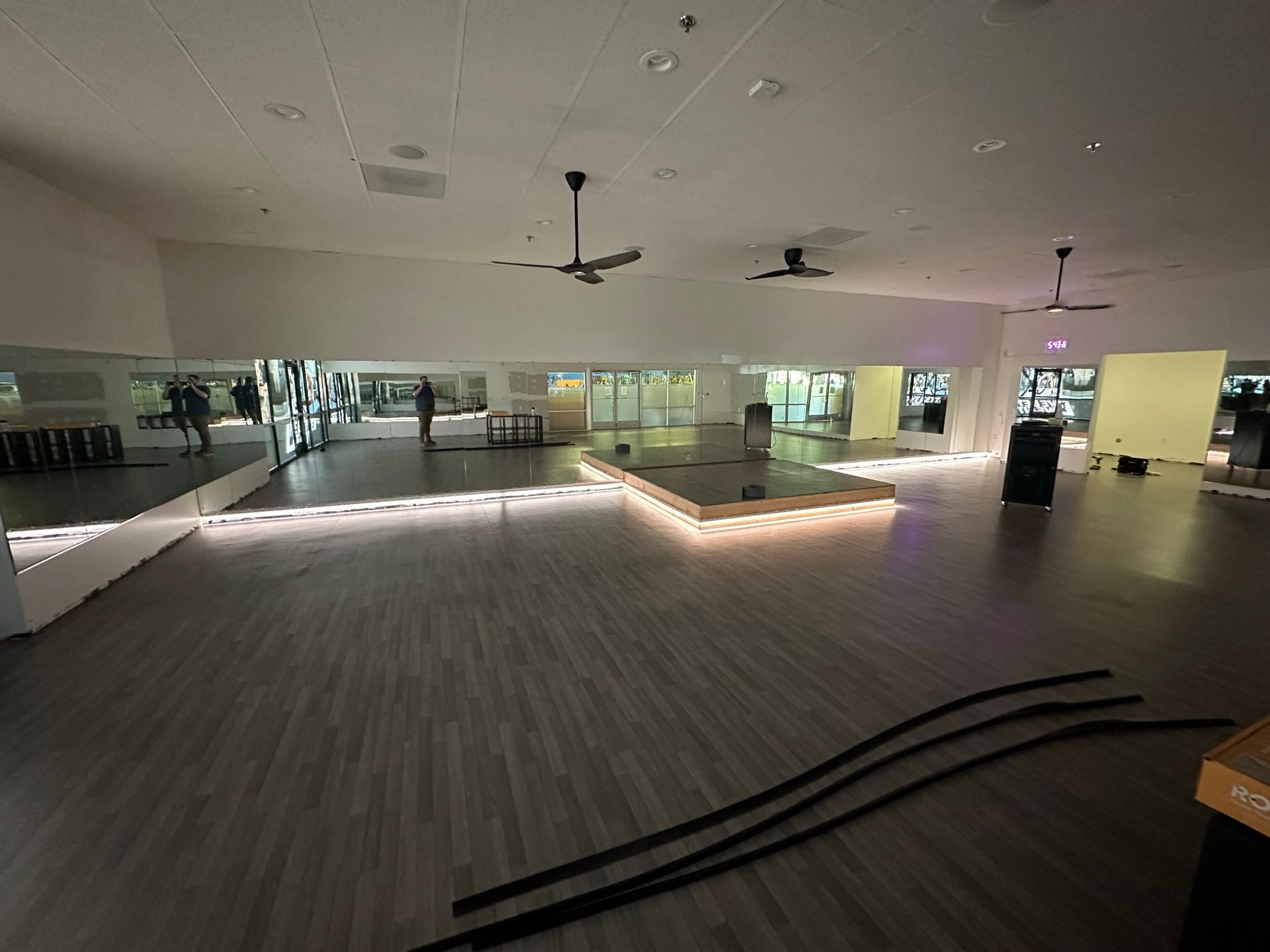 A dimly lit studio with a raised platform, wood-look flooring, ceiling fans, and a long mirrored wall.