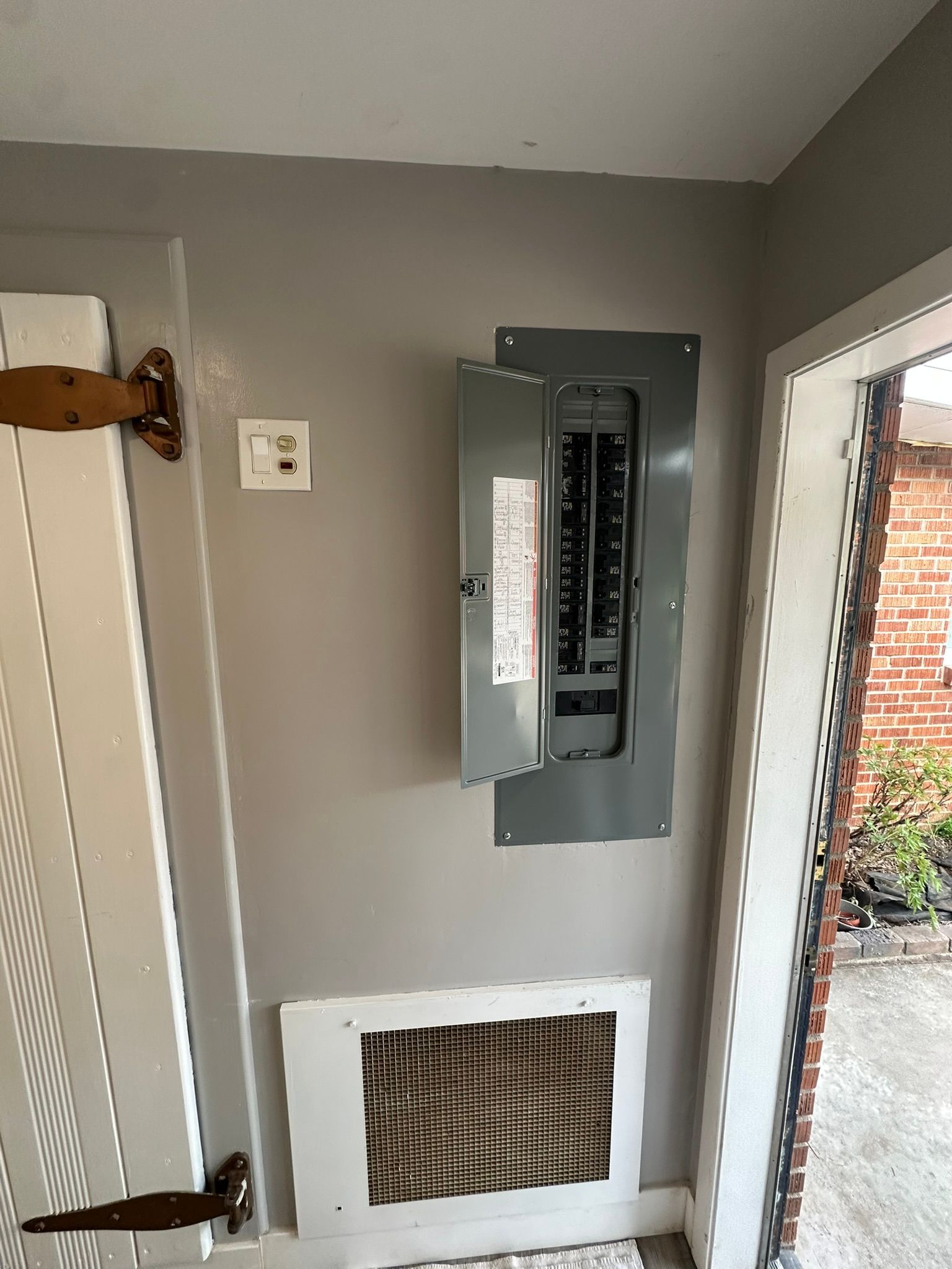 An electrical panel with an open door and a white HVAC filter intake grille mounted on a grey wall near a door frame.