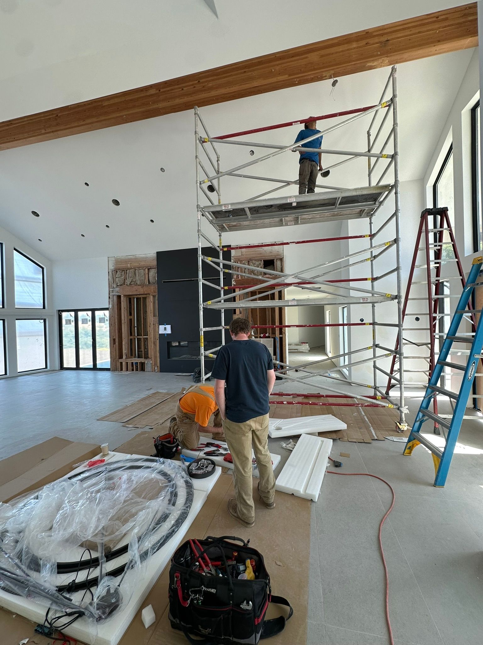 Two people working in a modern room under construction, one on high scaffolding and another standing near tools.