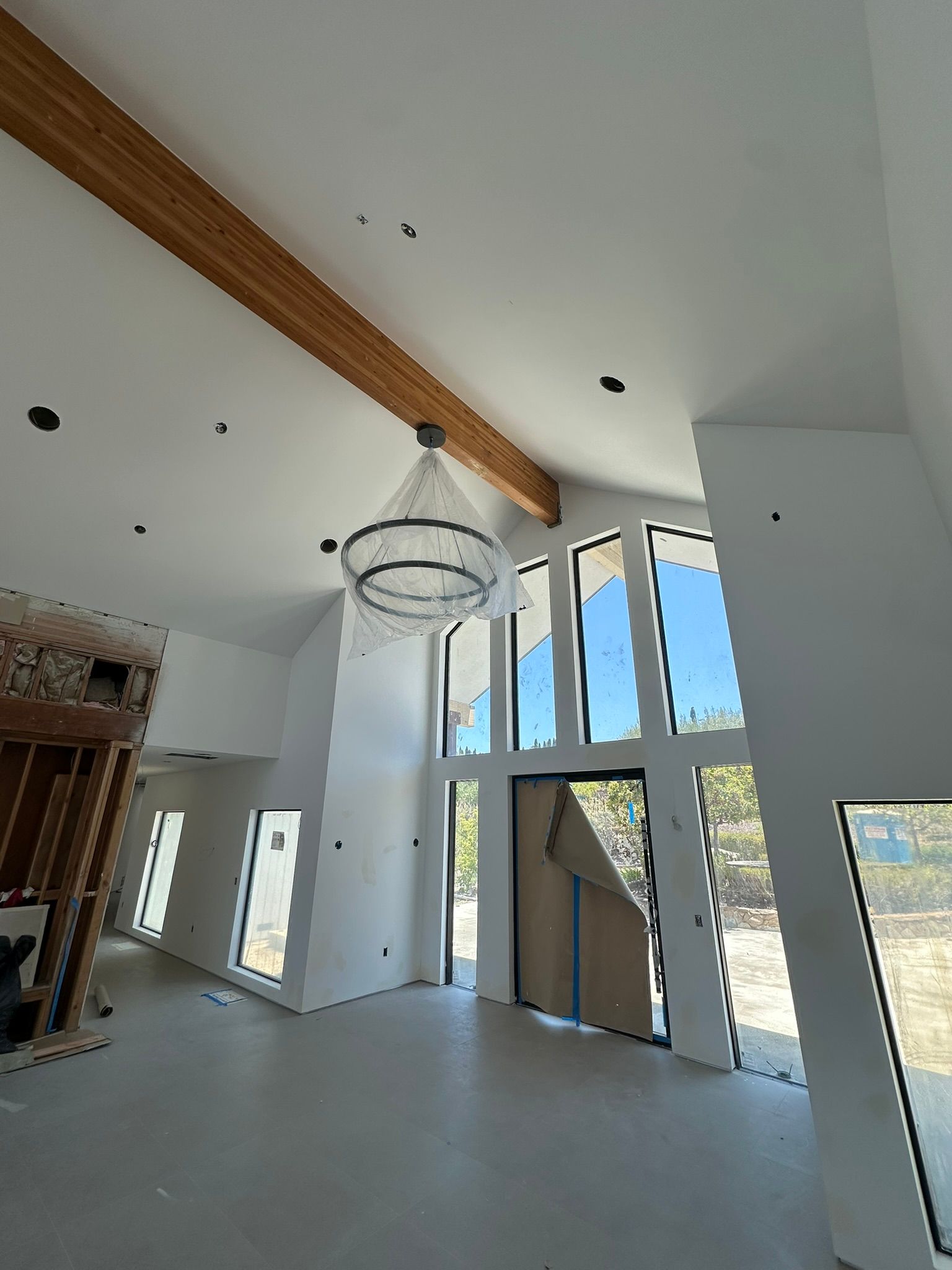 A brightly lit, modern room under construction with white walls, a large wooden beam, and floor-to-ceiling windows.