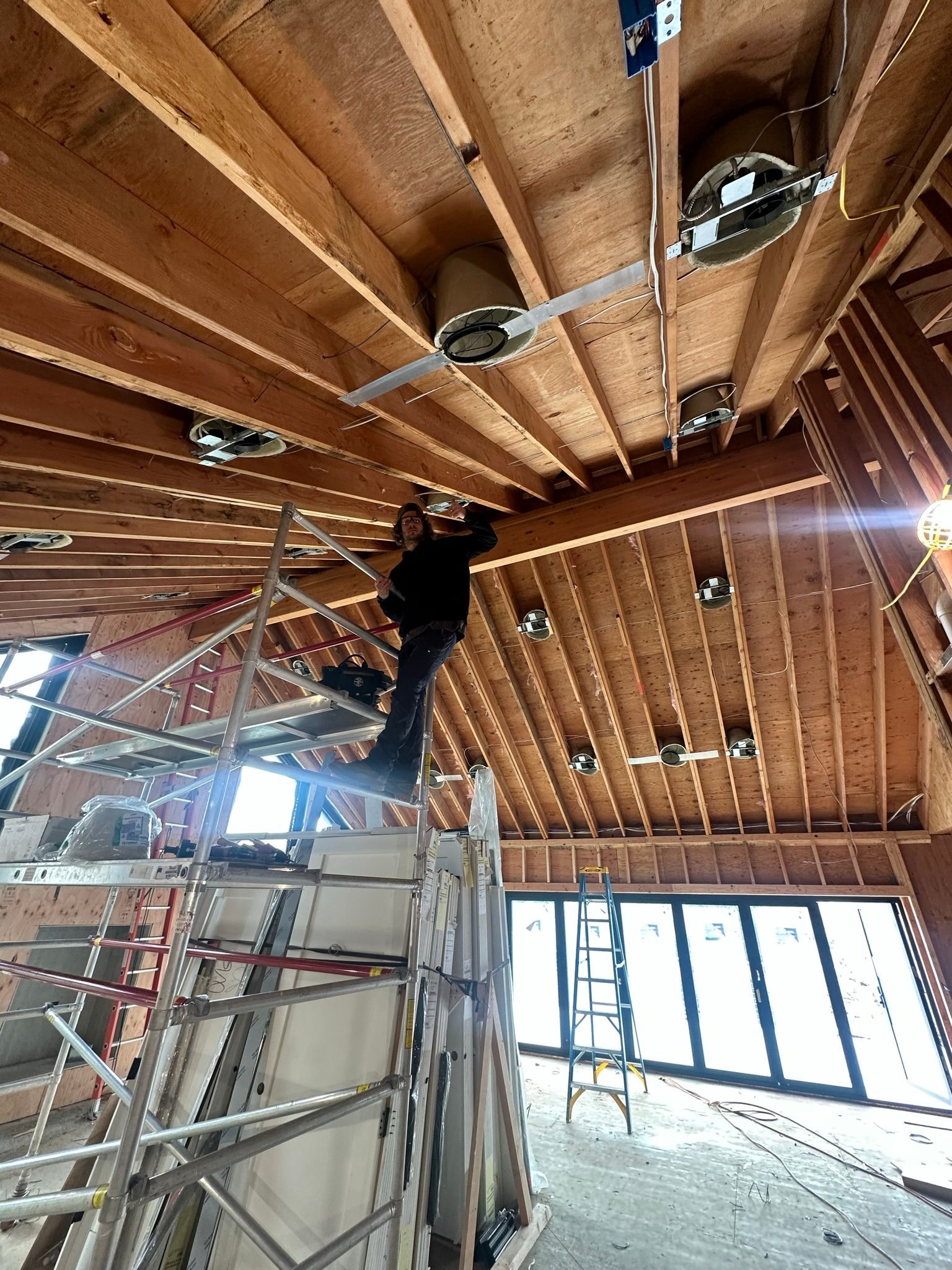 A worker on scaffolding installs recessed lighting in the wooden ceiling joists of a room under construction.