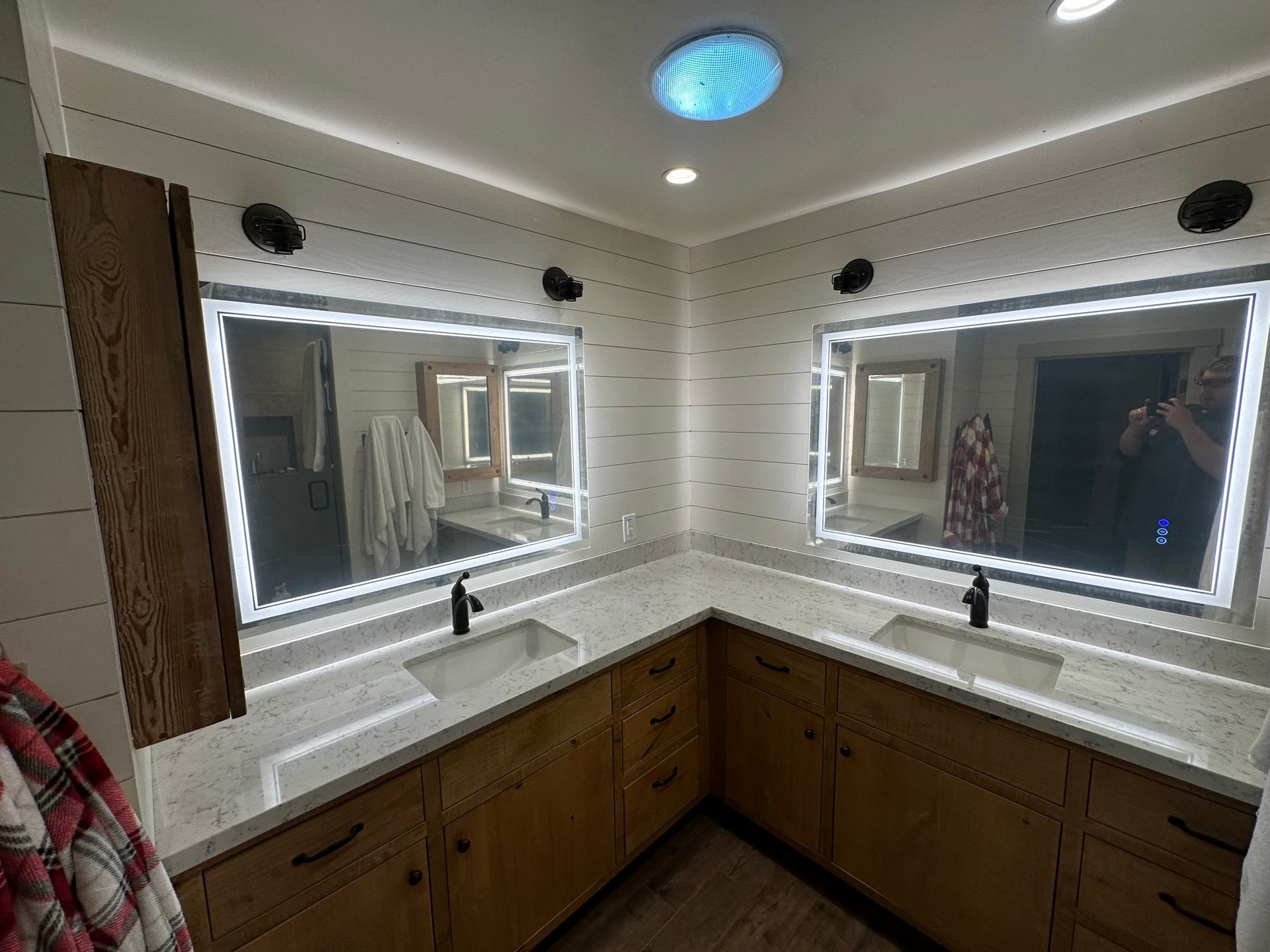 Double vanity bathroom with light wood cabinets, white countertops, two large illuminated mirrors, and shiplap walls.