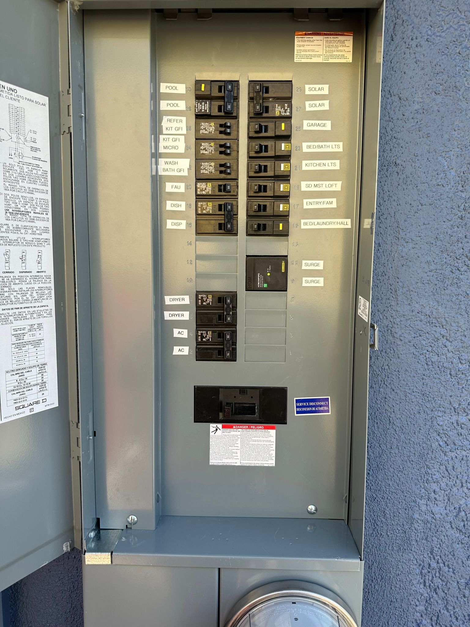 An open electrical breaker panel mounted on an exterior grey stucco wall with labels next to each switch.