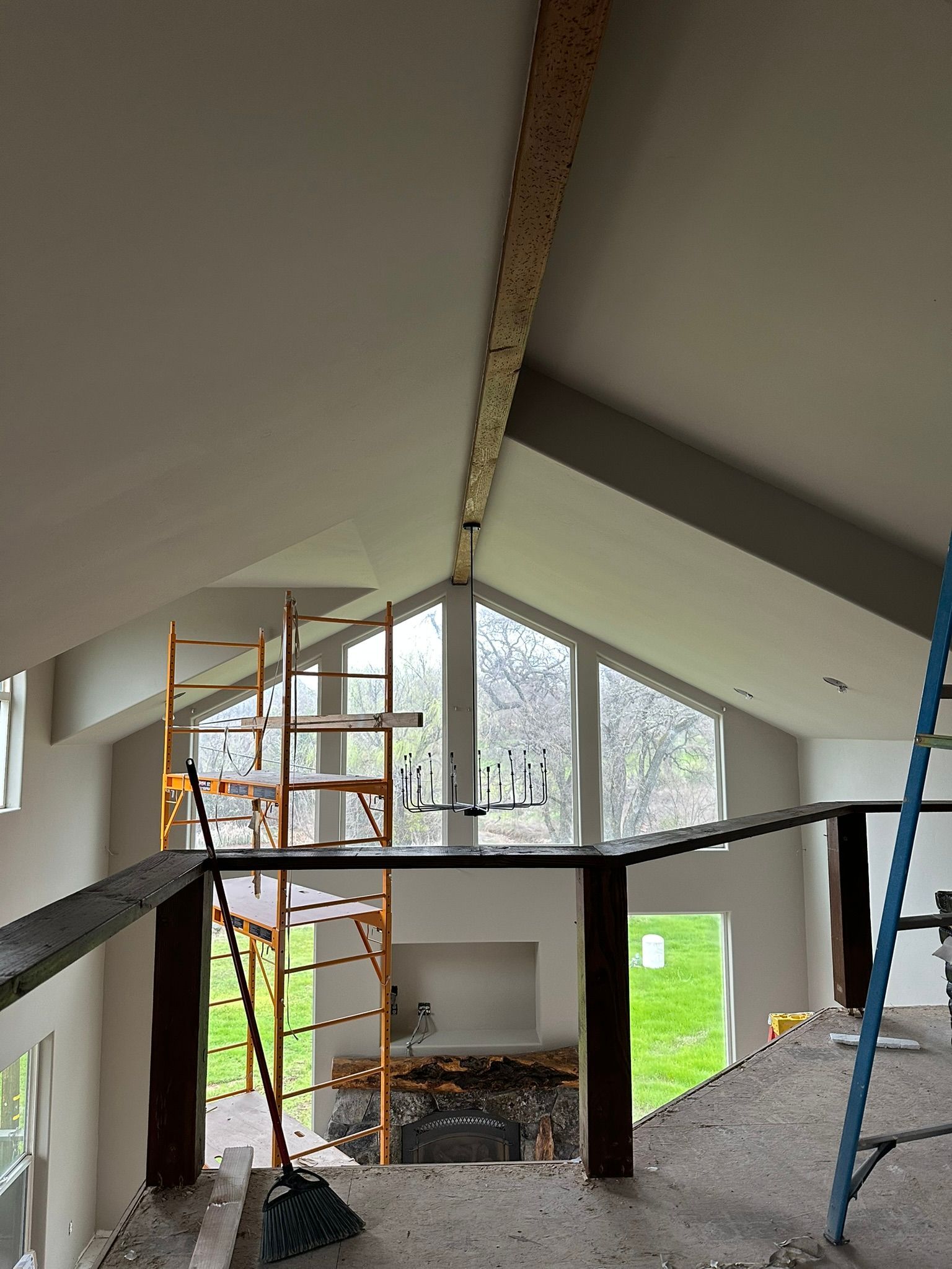 A room under construction with a high vaulted ceiling, exposed wooden beam, large window, and metal scaffolding.