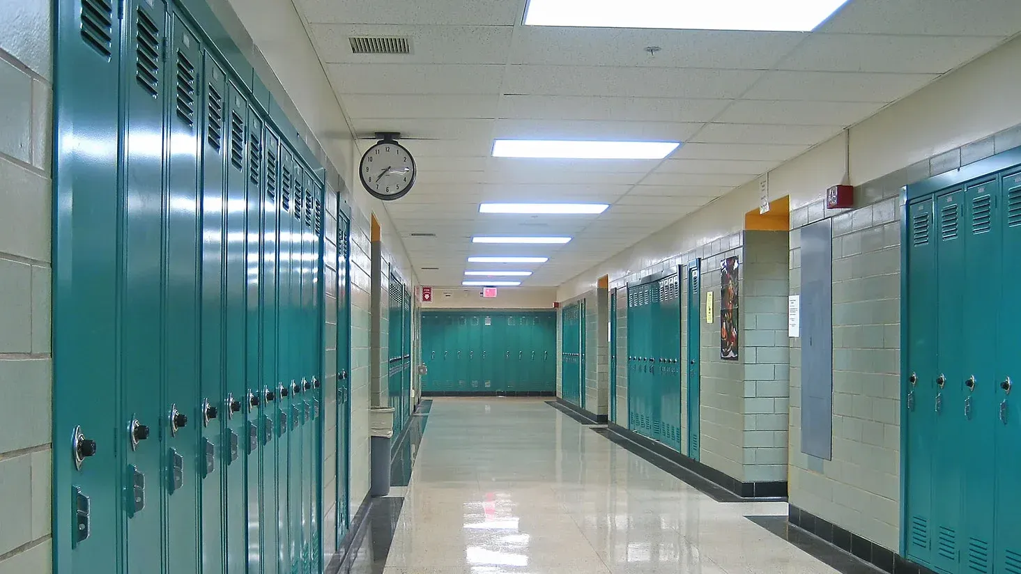 An empty school hallway with teal lockers lining both sides, polished tile floors, and overhead fluorescent lights.
