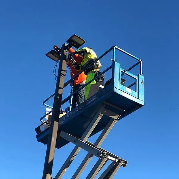 Two workers in high-visibility gear stand in a blue scissor lift, repairing lighting atop a pole against a clear blue sky.