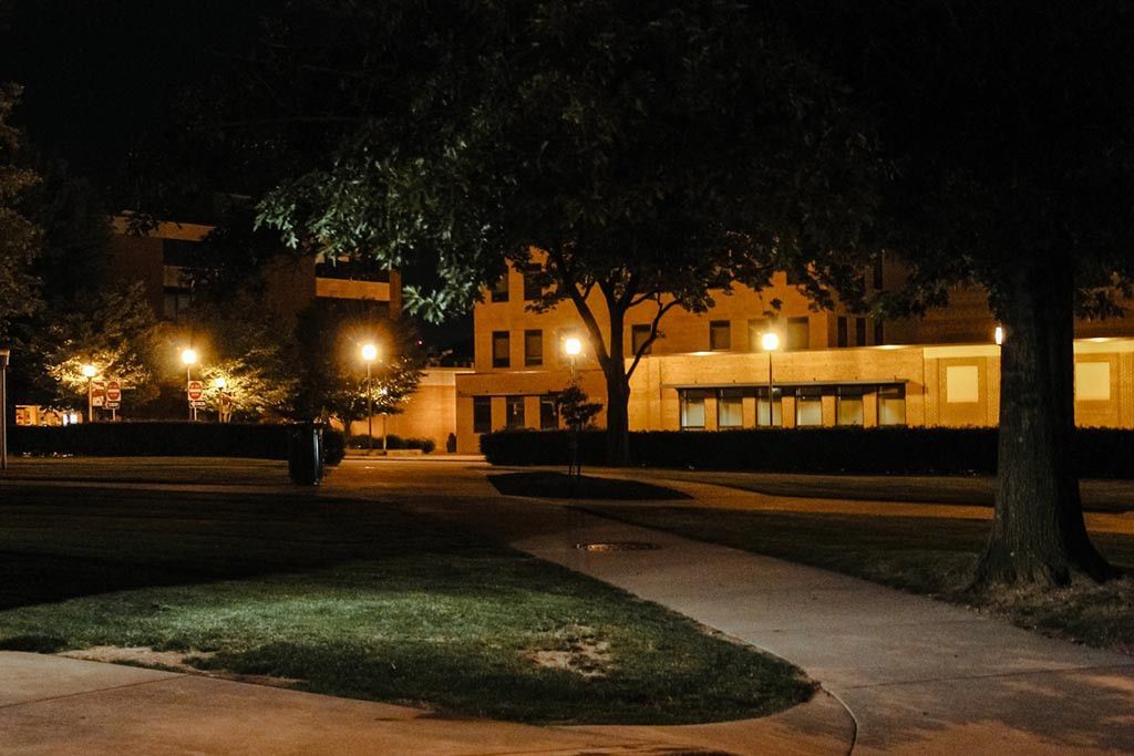A night scene showing a paved walkway leading toward a lit, multi-story building through a park with trees.