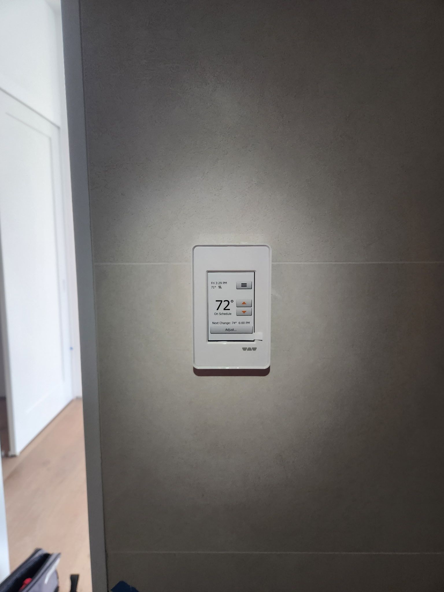 A white wall-mounted thermostat control panel with a digital display, centered on a gray wall.