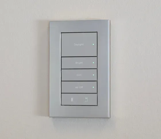 A silver, rectangular wall-mounted light control panel with five backlit buttons labeled with text and icons.