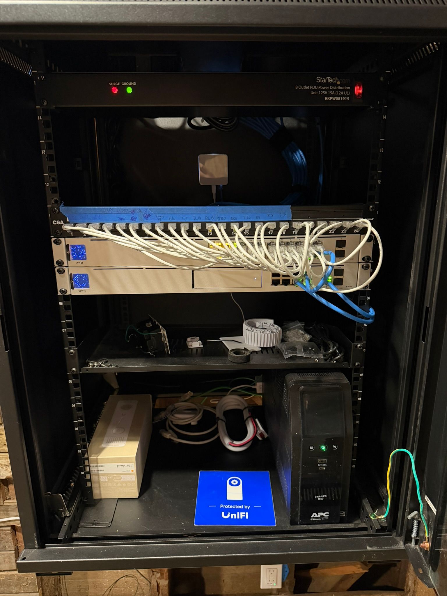 A server rack containing network equipment, cables, and a UPS on a bottom shelf within a black metal enclosure.