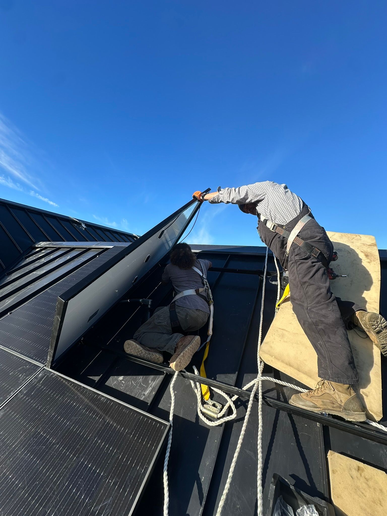 Two workers in safety harnesses install a solar panel on a black standing-seam metal roof under a bright blue sky.