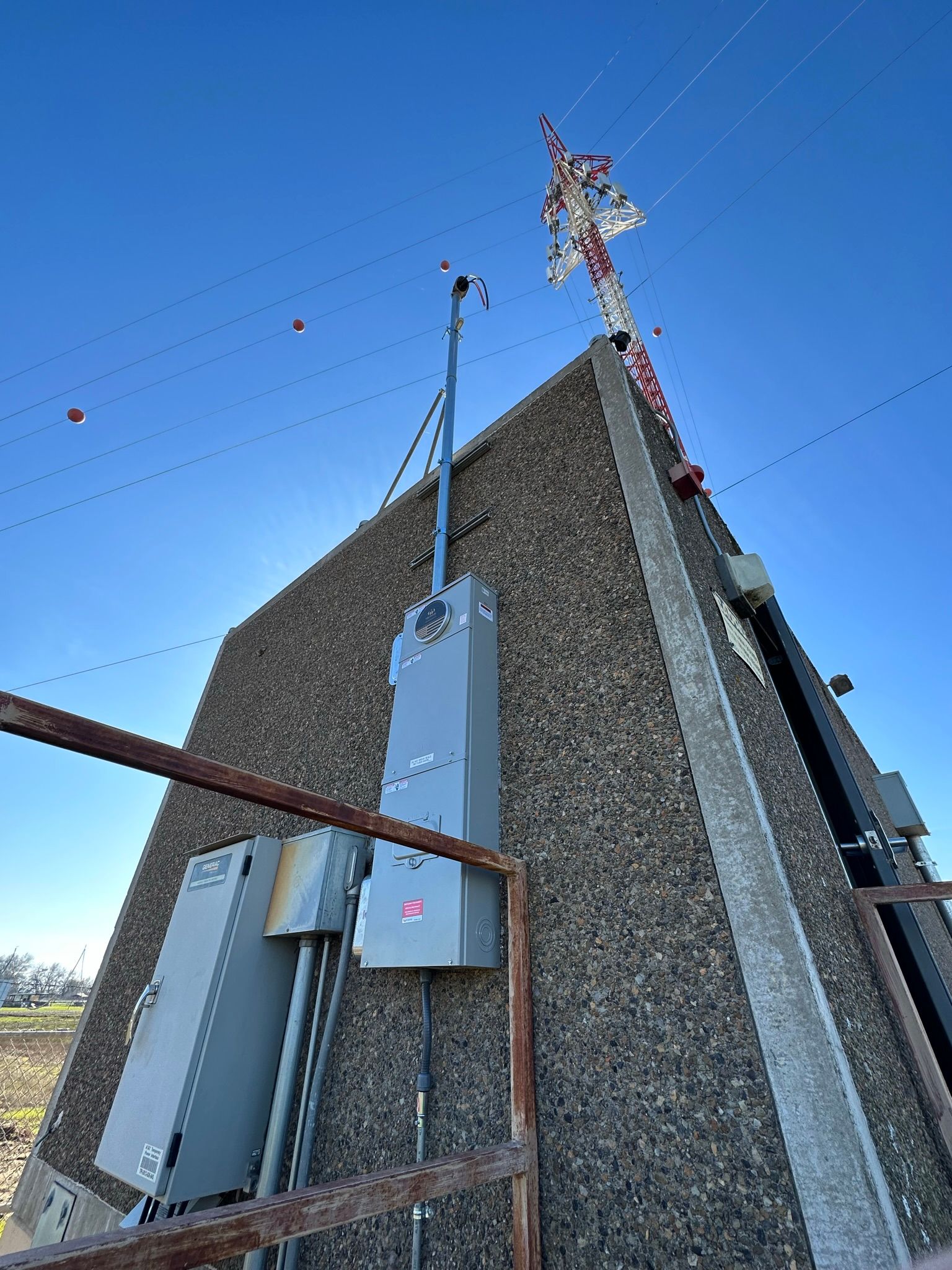An upward angle view of grey electrical panels mounted on a pebble-dash wall with a tall utility tower against a blue sky.