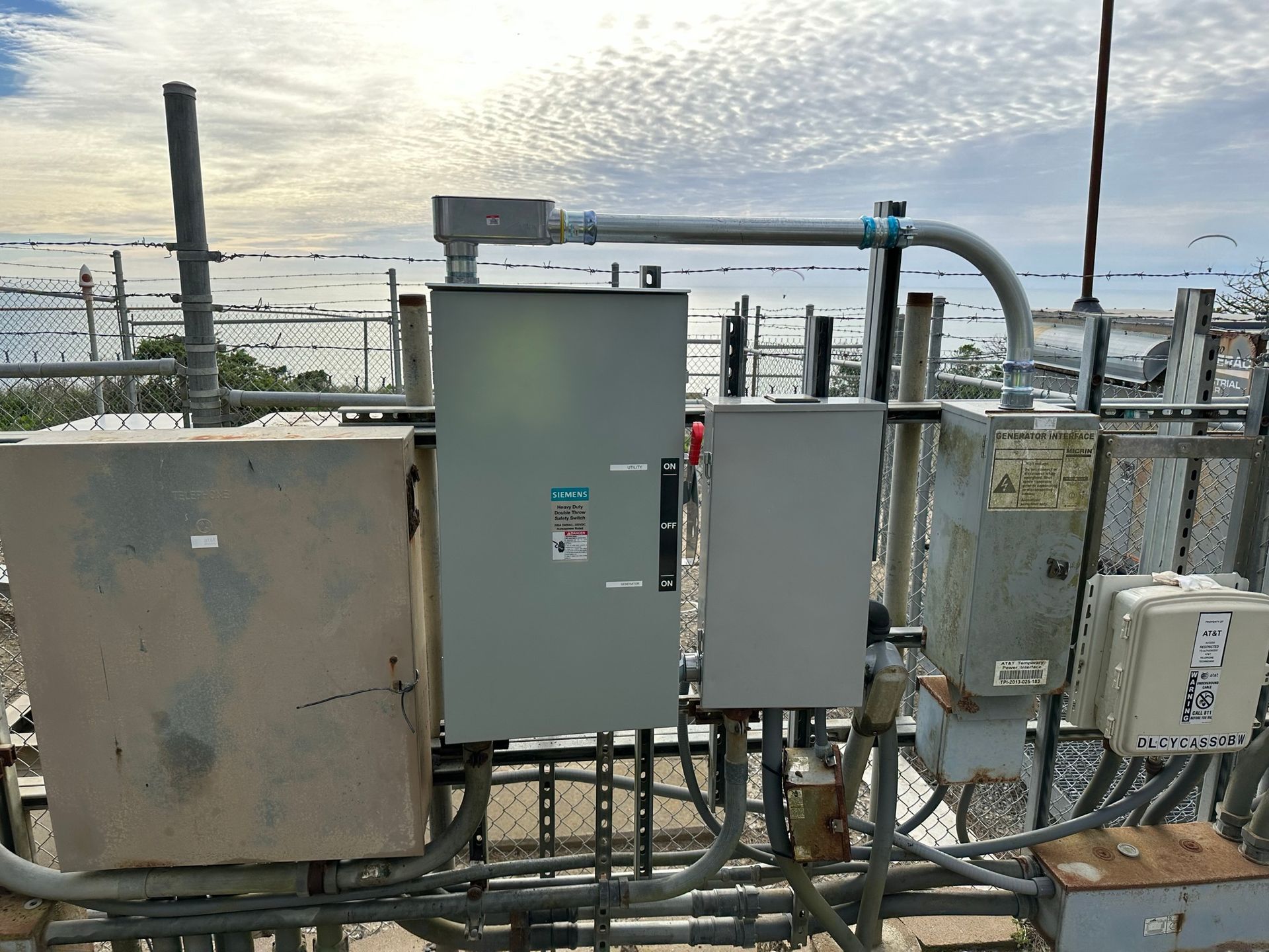 A series of metal electrical boxes and conduits mounted on a rack against a backdrop of a cloudy sky and distant water.