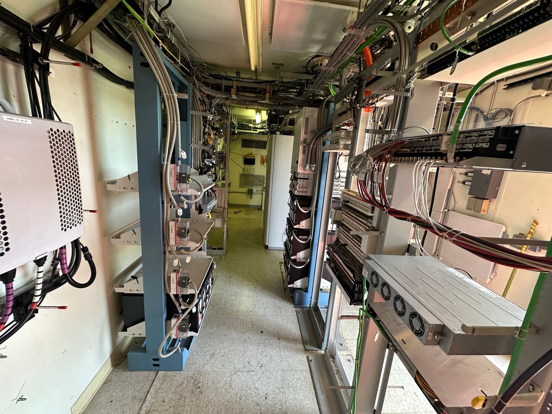 A narrow technical room containing server racks, complex wiring, and communication equipment lining the walls.