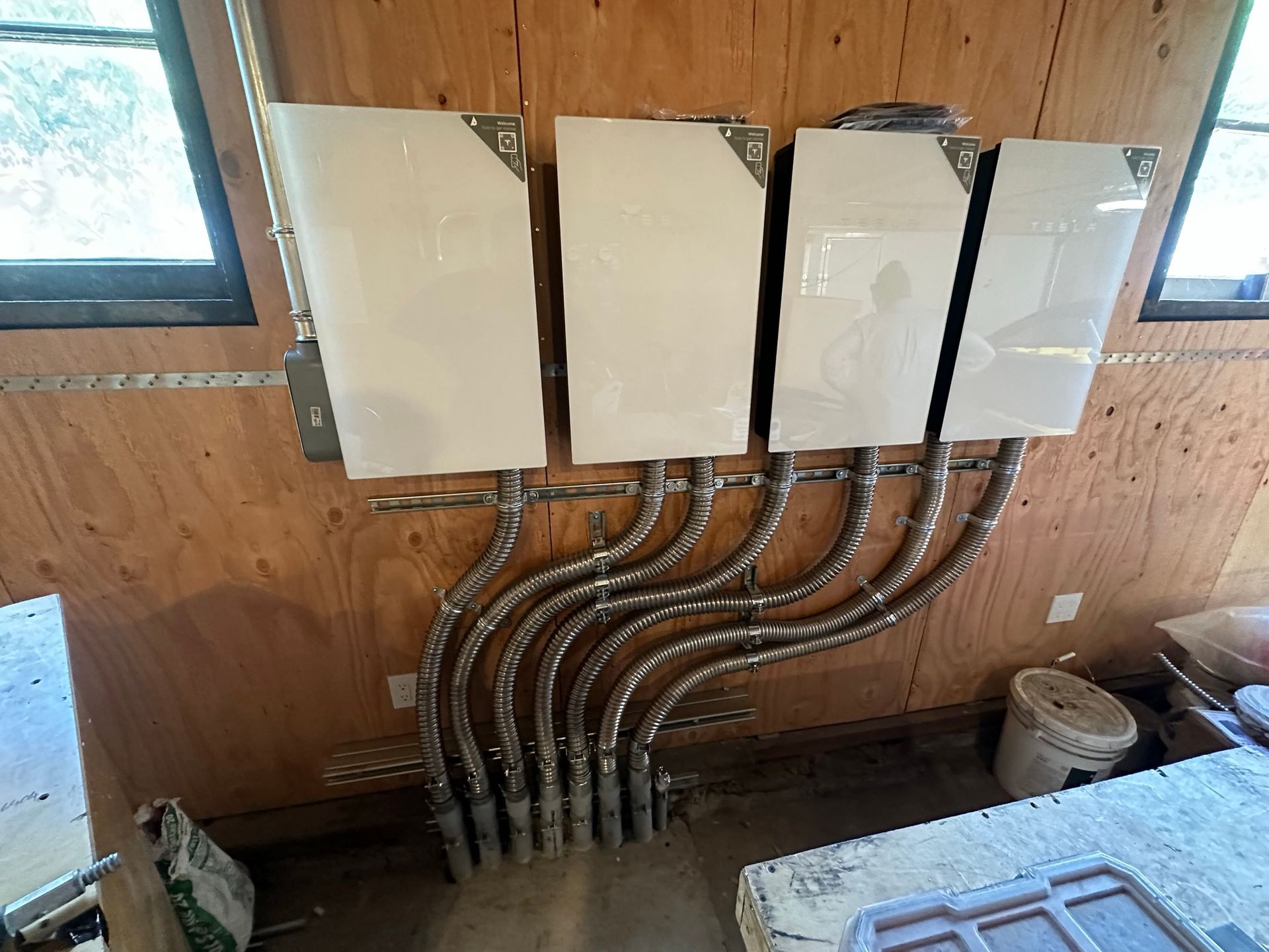Four white energy storage units mounted on a wooden wall, connected by silver flexible conduits running to the floor.