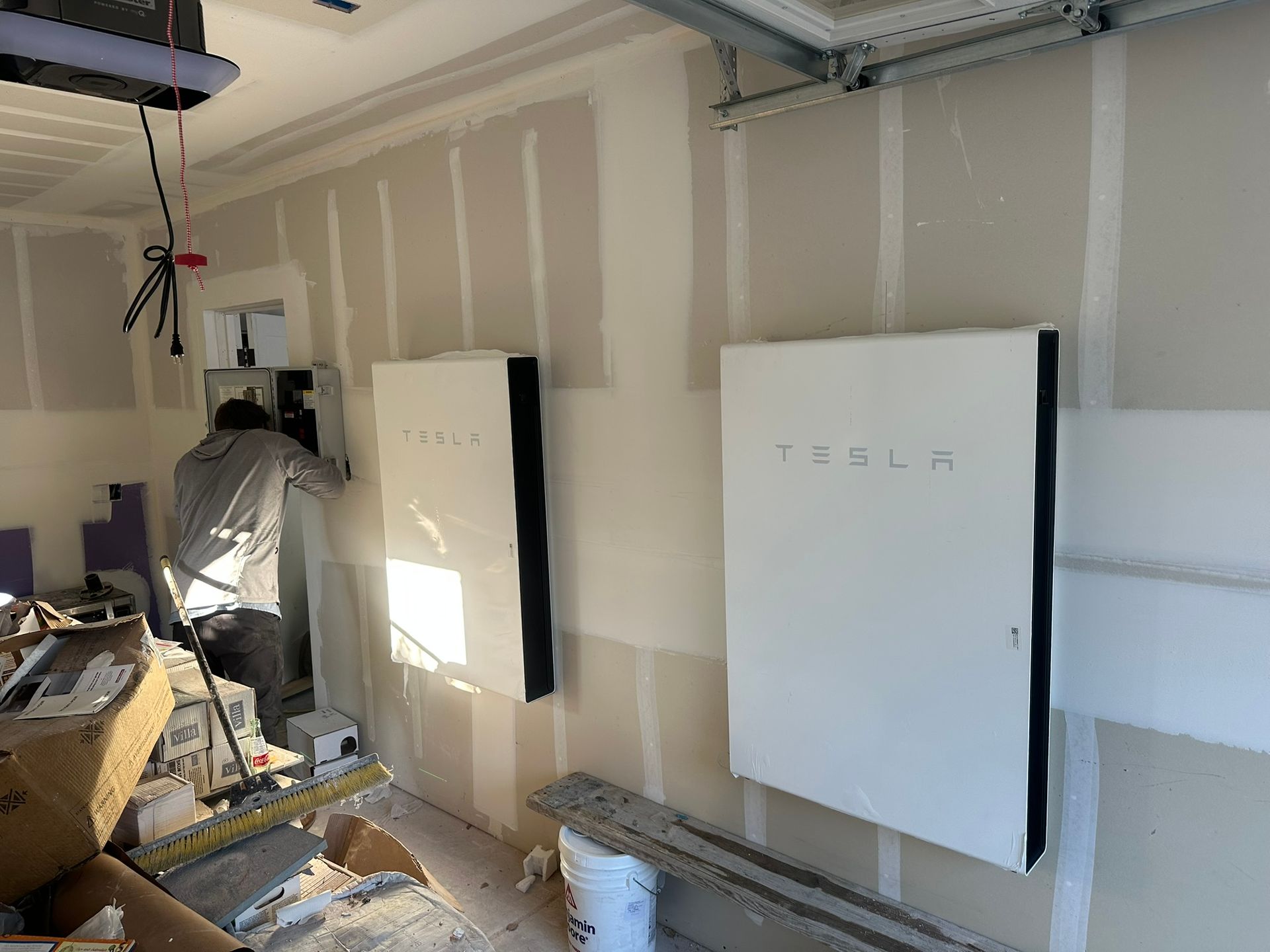 A technician installs two white Tesla Powerwalls on a garage wall with unfinished drywall.