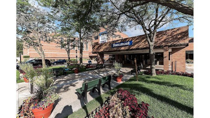 A large brick building with a blue sign on it is surrounded by trees and flowers.