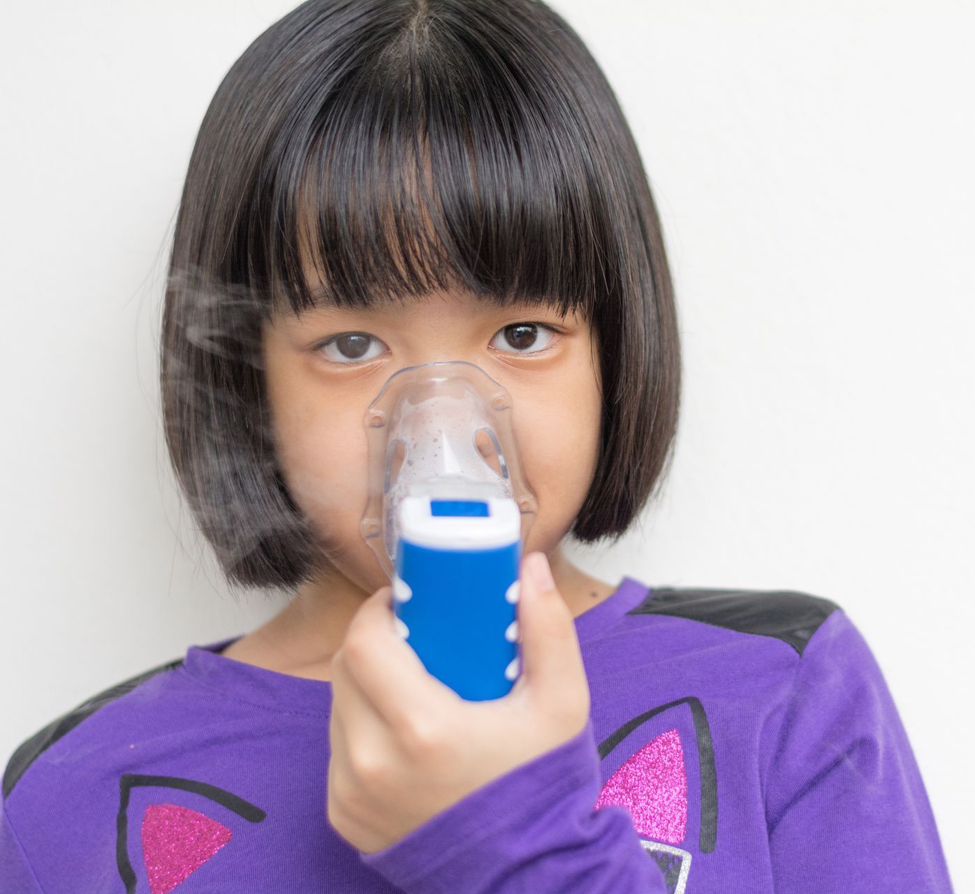 A young Asian girl with short black hair wearing a purple shirt uses a nebulizer, inhaling mist.