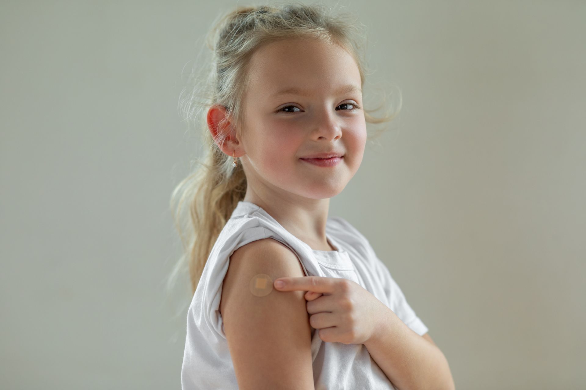 A young girl with blonde hair points to a bandage on her upper arm, smiling at the camera. She wears a white t-shirt.