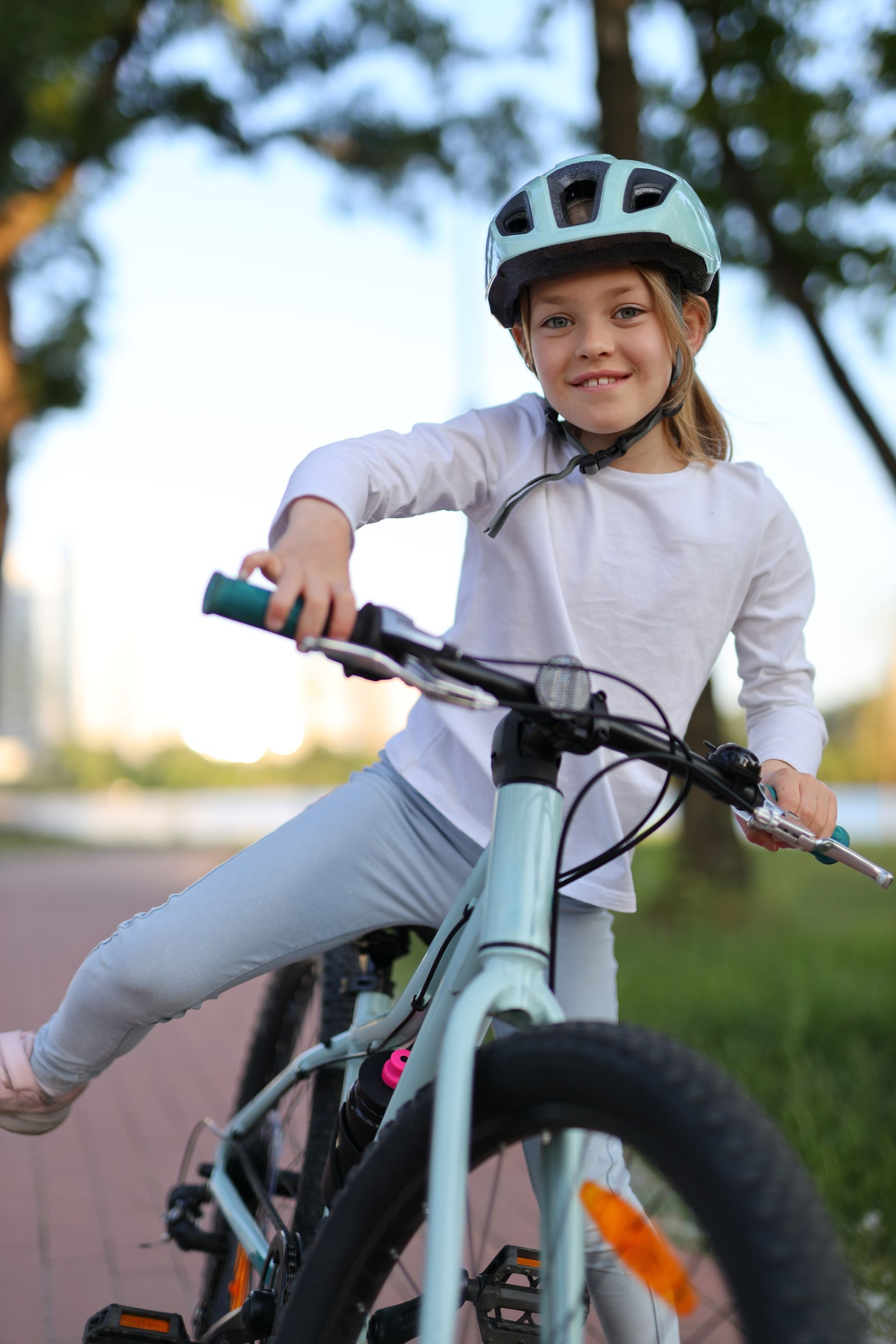 Girl in a blue helmet and bike leans sideways on a blue bicycle on a paved path, smiling at the camera.