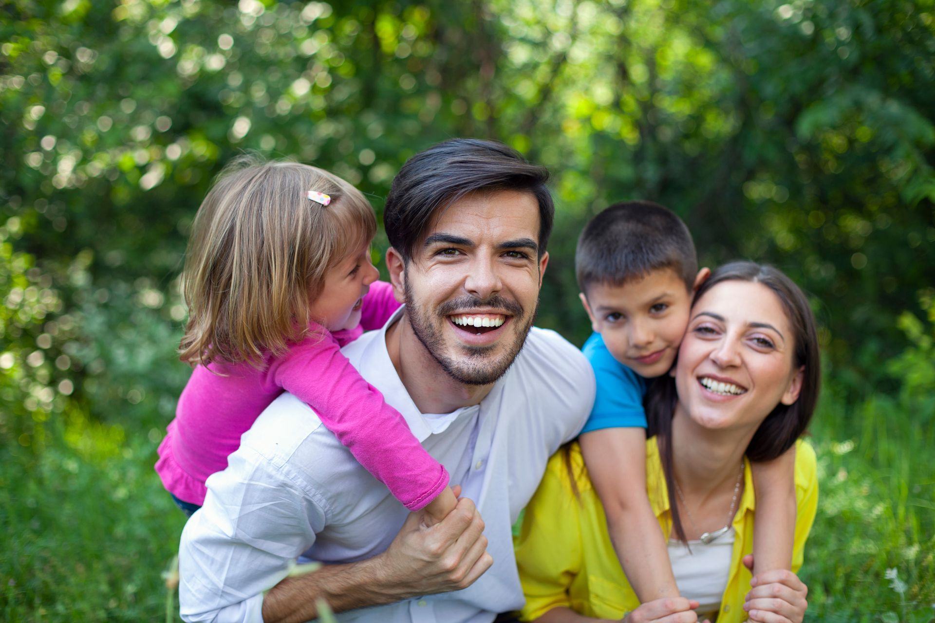 Family of four smiling and hugging outdoors. Two children are perched on the parents' backs, surrounded by greenery.