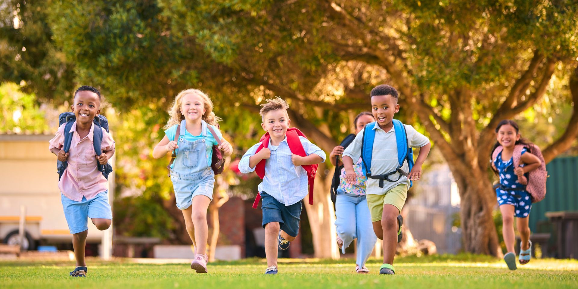 A group of diverse children with backpacks run joyfully on a grassy field. A large tree provides shade.