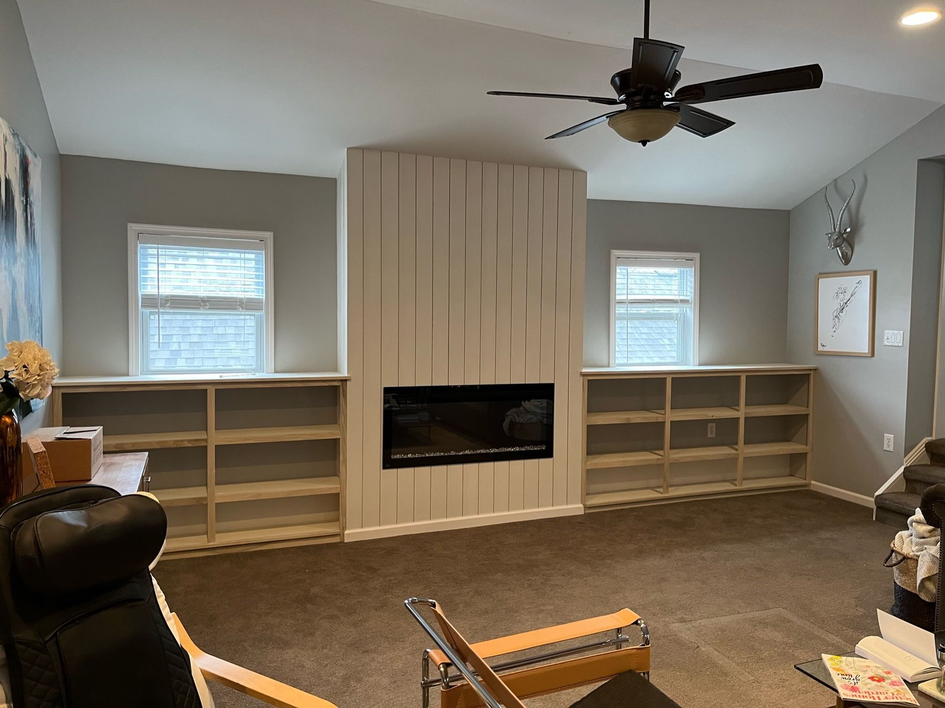 Living room with fireplace, built-in shelves, windows. Light-colored walls and brown carpet. Black ceiling fan.