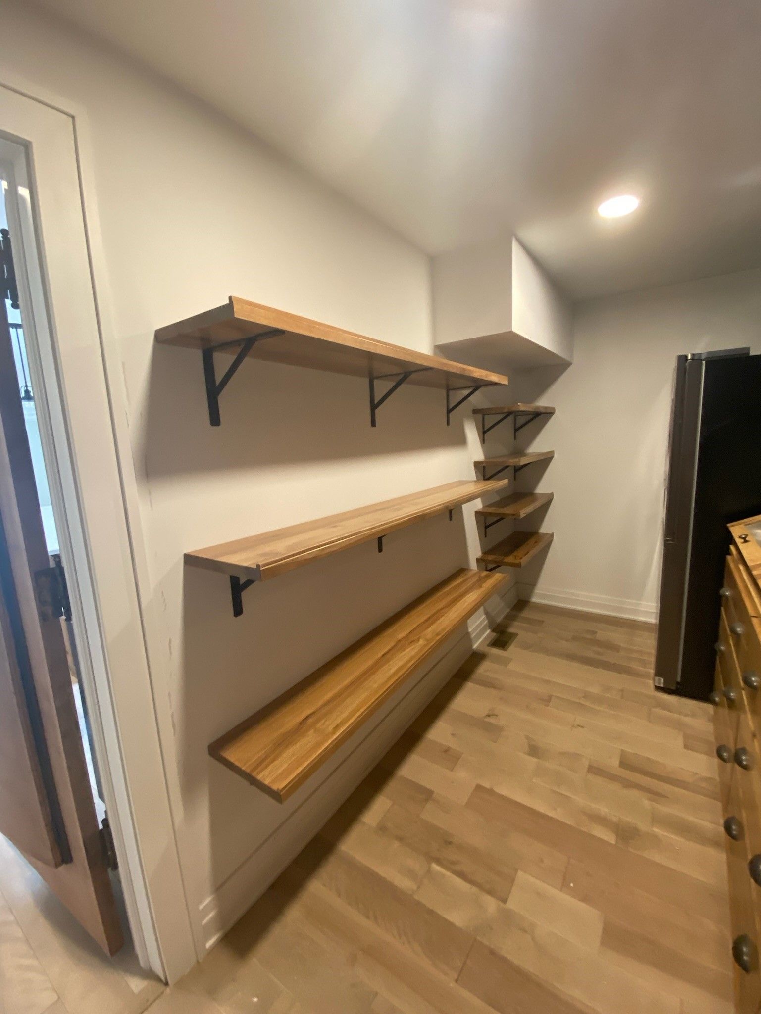 Wooden shelves on a white wall in a kitchen. Light wood flooring and a black refrigerator.