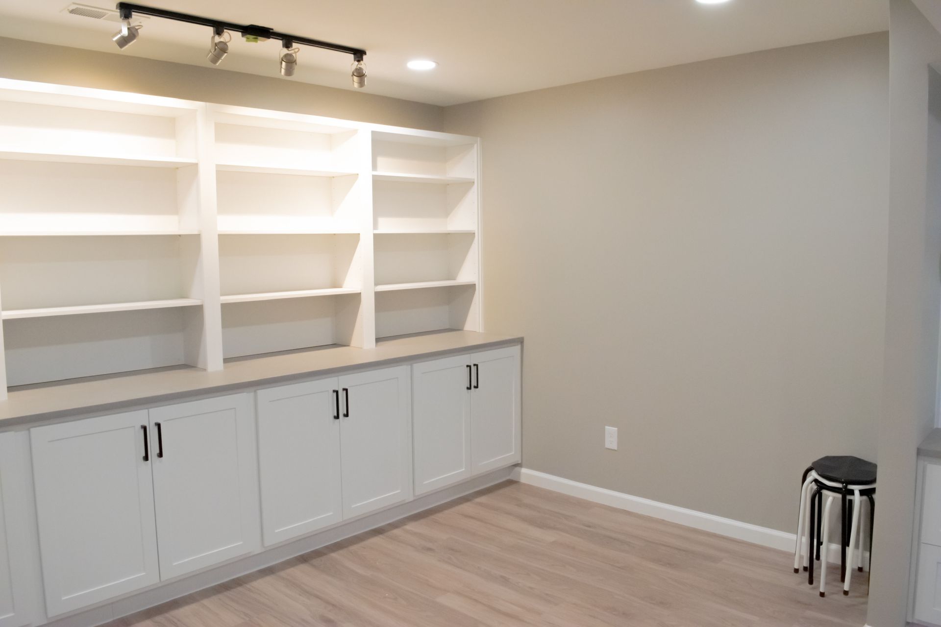 Built-in white bookshelves and cabinets in a room with light wood floors and gray walls; a stool sits nearby.