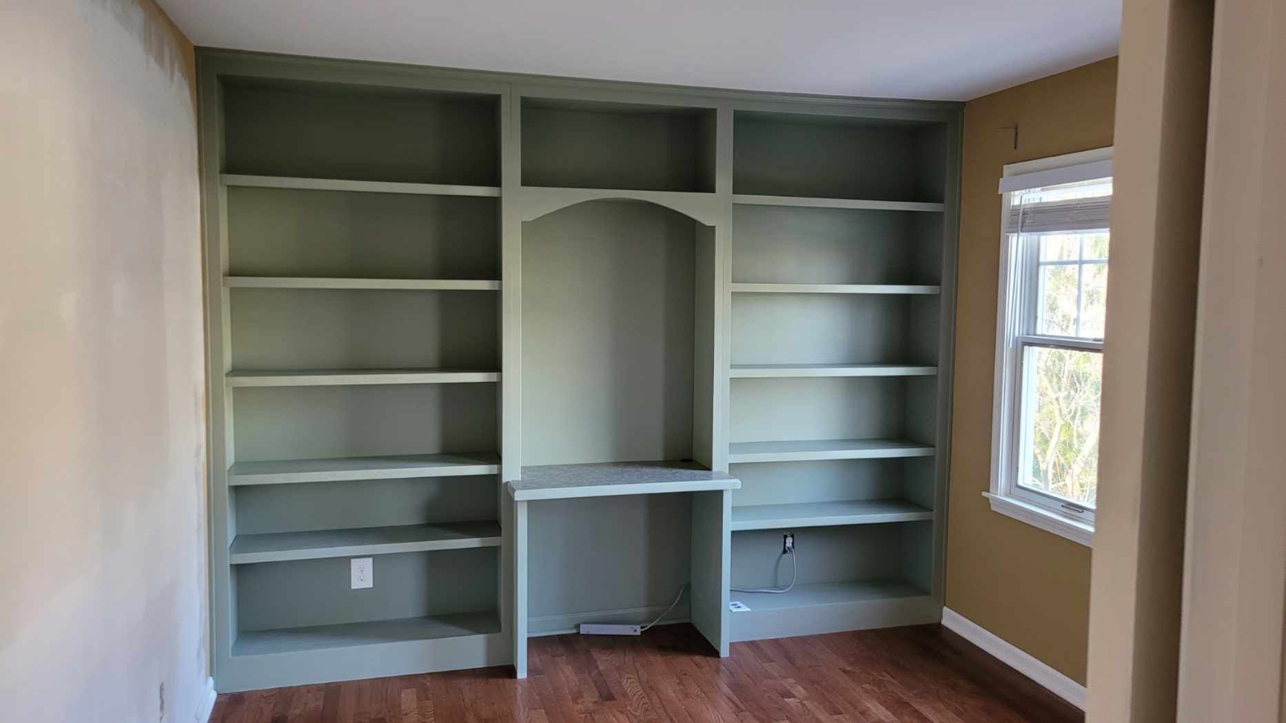 Built-in green bookshelves with a desk area, set in a room with a window and hardwood floor.