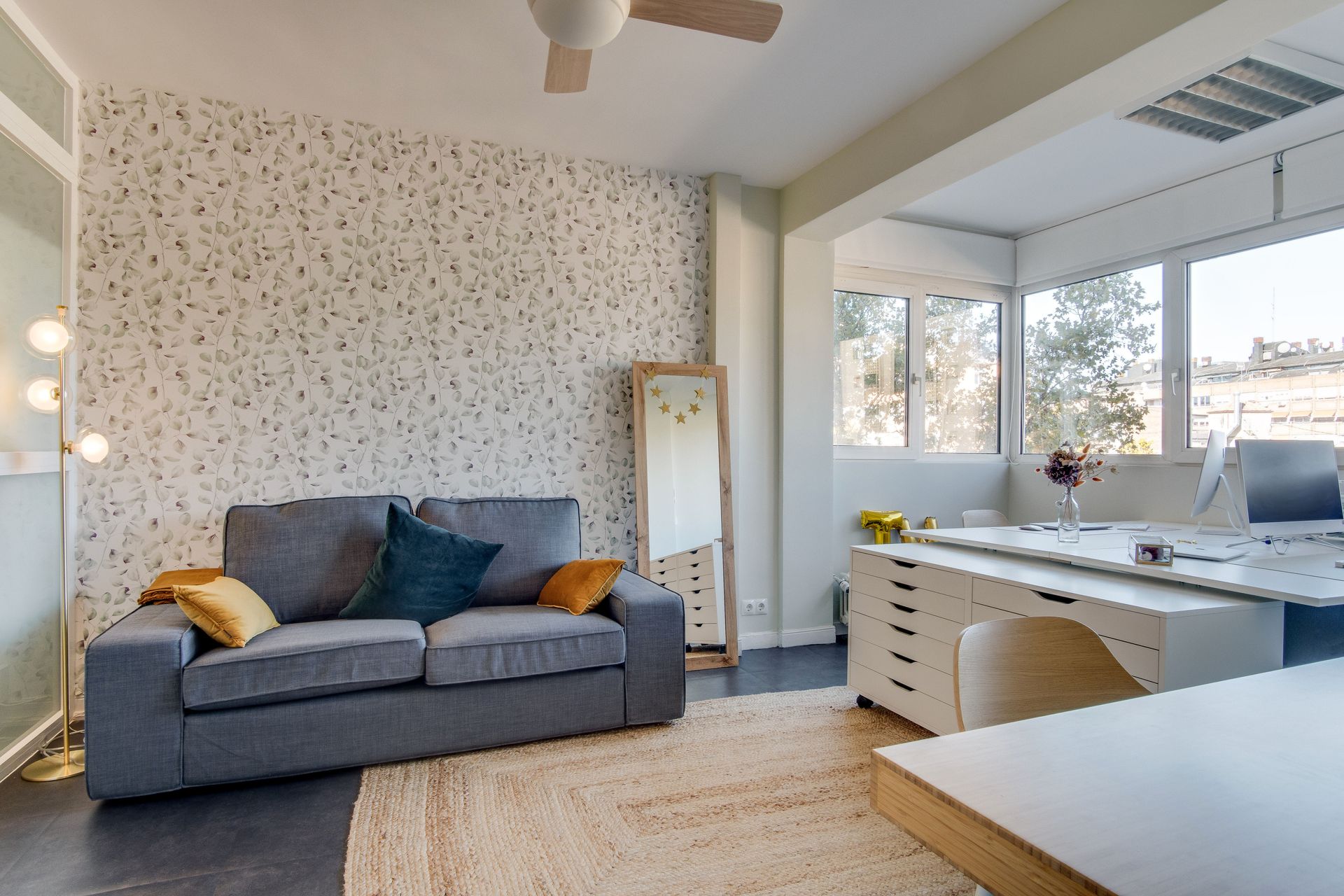 Cozy living room with gray couch, patterned wallpaper, rug, and a desk area with windows.