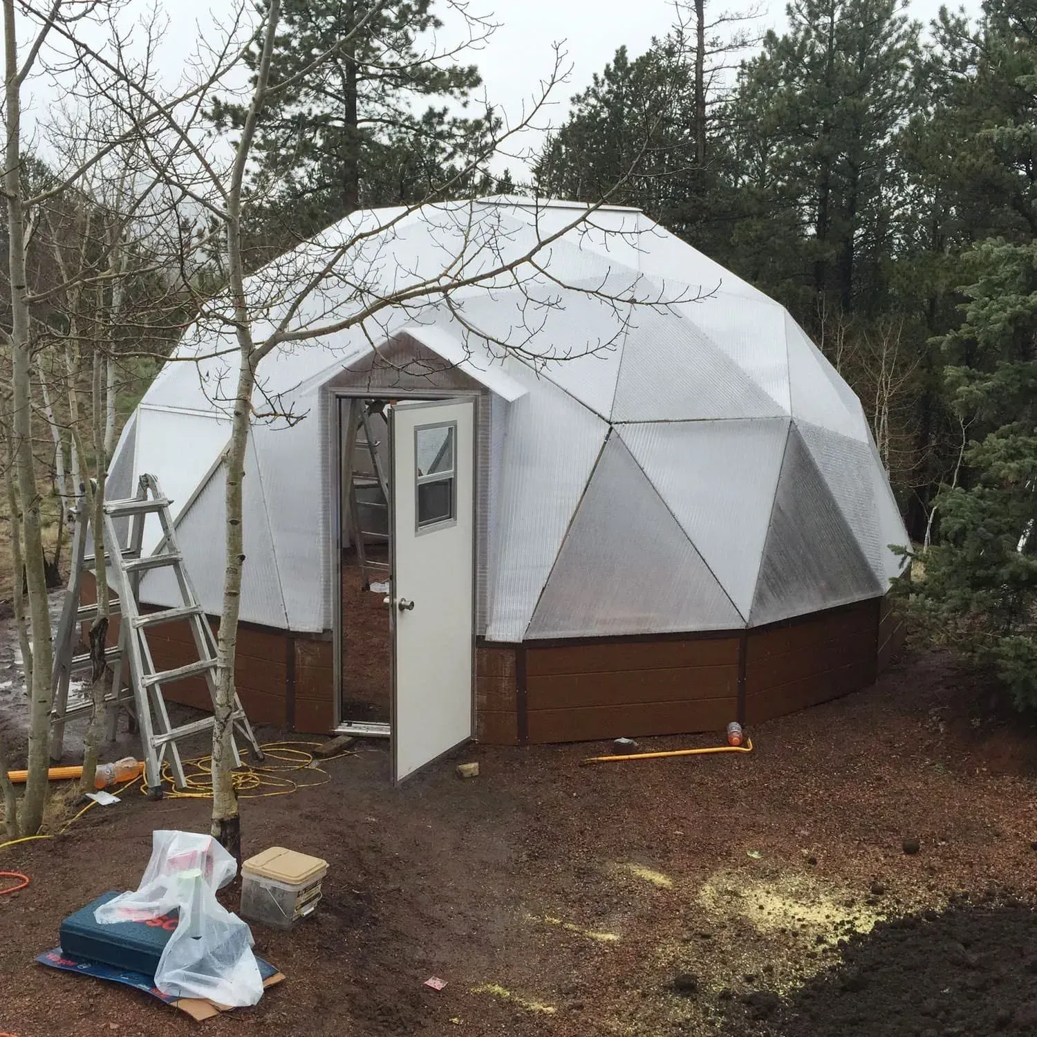 Geodesic dome greenhouse with an open door, brown base, and clear panels, outdoors.