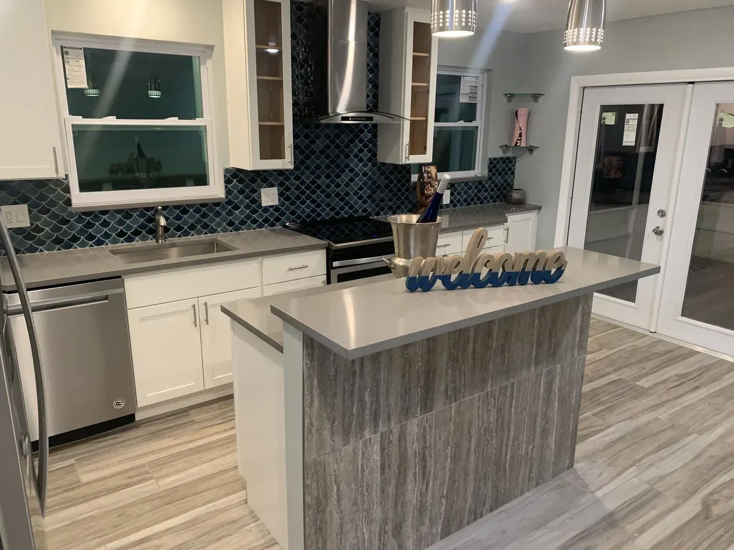 Modern kitchen with white cabinets, blue backsplash, and light wood flooring. Island with a gray countertop.