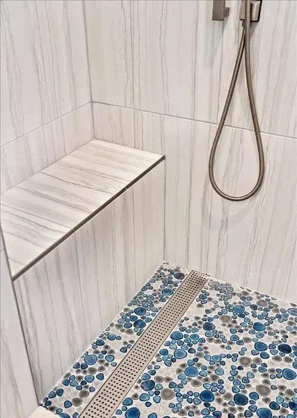Shower with white marble-look tile, a bench, and blue pebble floor with linear drain and brushed metal fixtures.