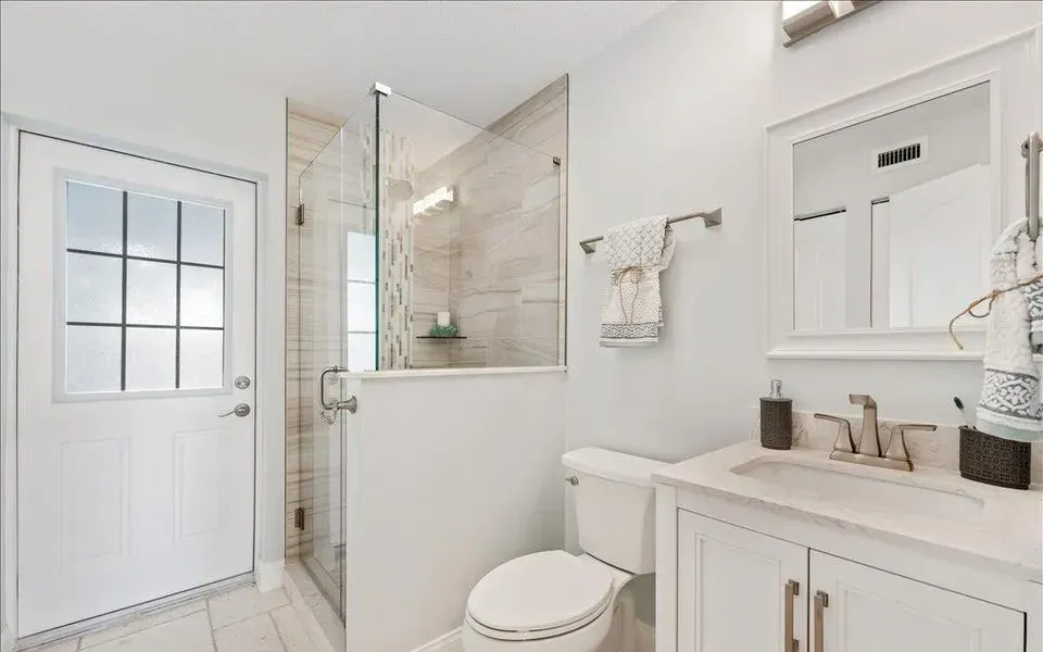 Bright bathroom with white walls, shower with glass door, toilet, sink, and door to outside.