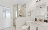 Bright bathroom with white walls, shower with glass door, toilet, sink, and door to outside.